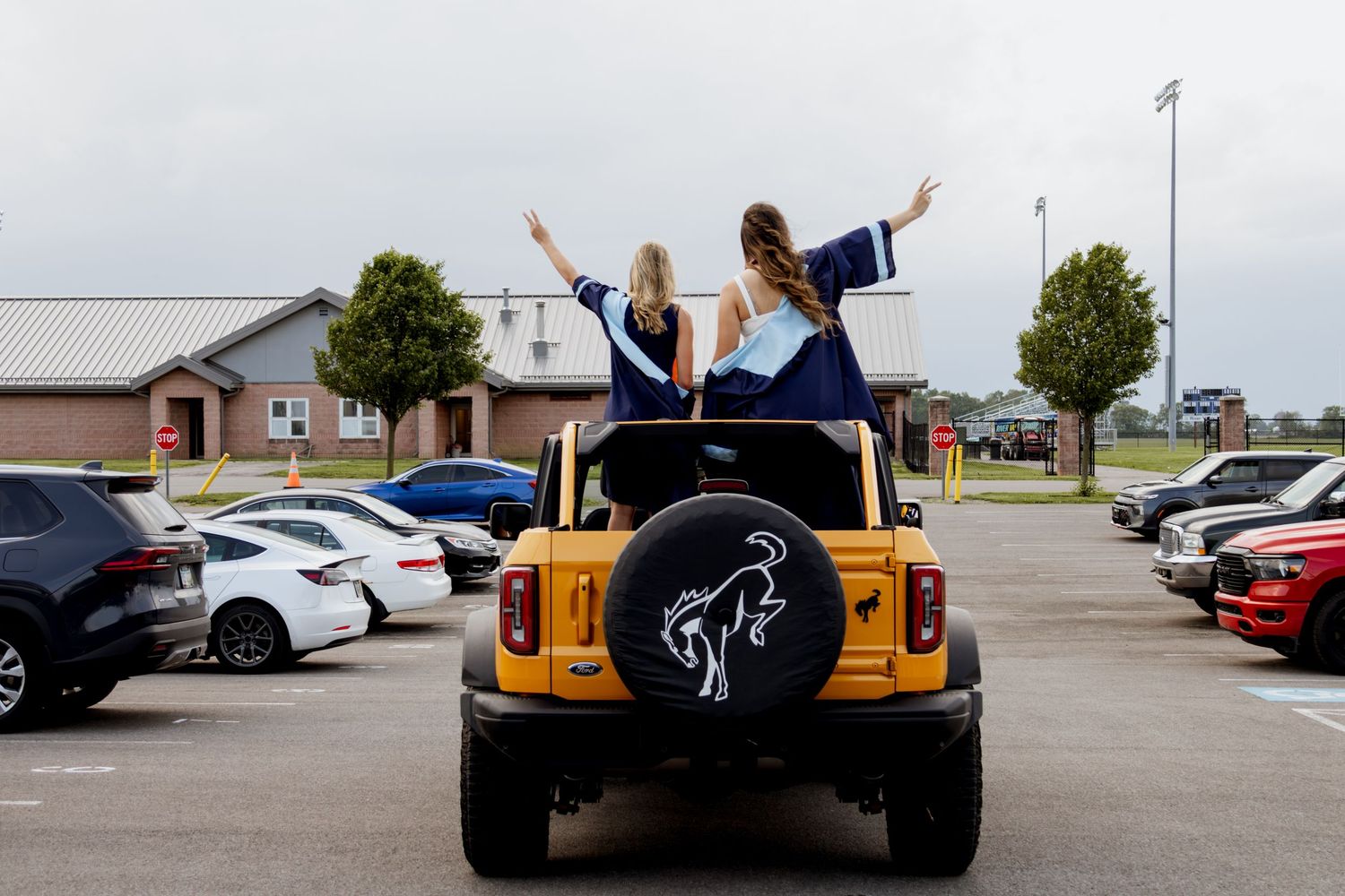 Two graduates in caps and gowns stand in a yellow Bronco in a parking lot, arms raised in celebration.