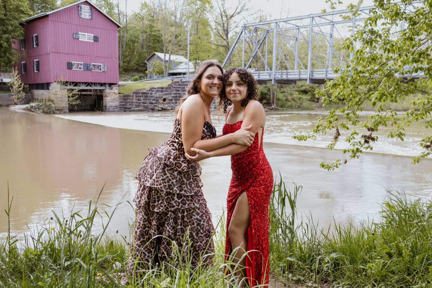 Two young women in formal gowns pose together near a river with a red barn backdrop.