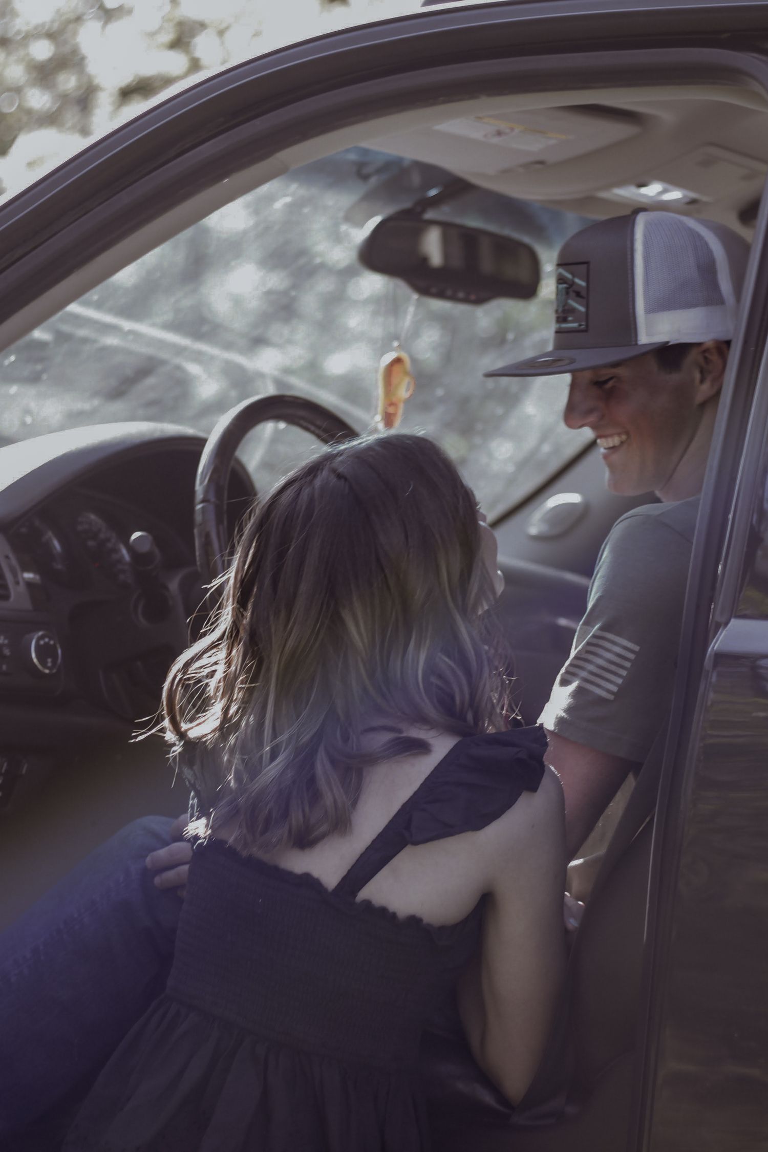A young couple shares a candid, smiling moment beside an open truck door in dappled sunlight.