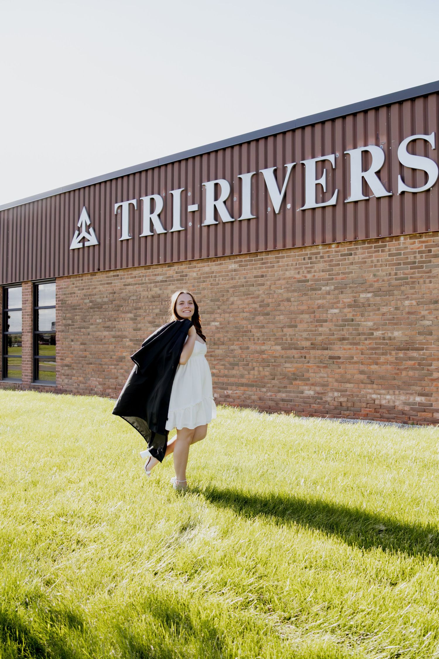Young woman in white dress holding graduation gown outside Tri-Rivers school building.