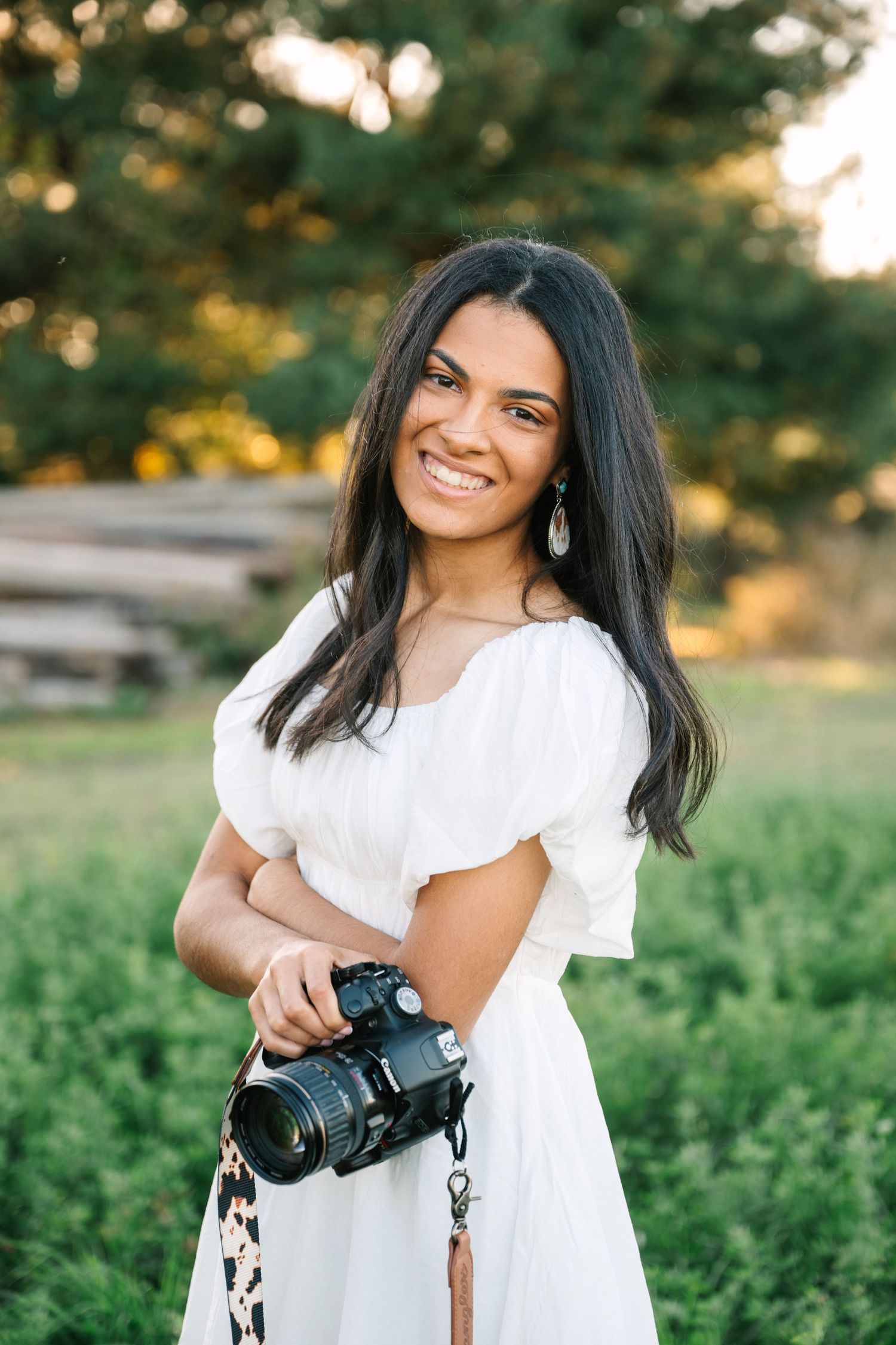 Smiling young woman in white puff-sleeve dress holding a DSLR camera in a sunny green field.