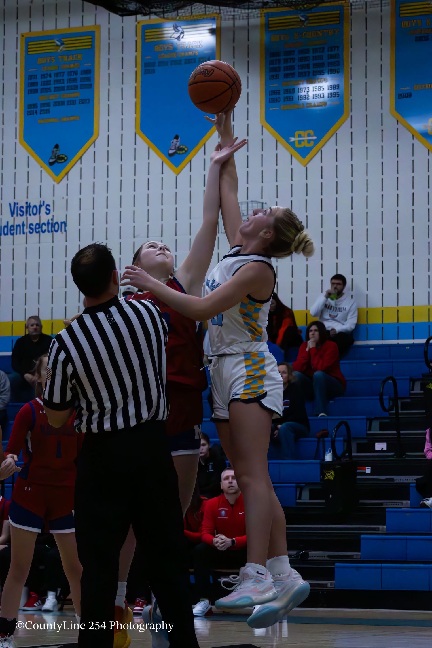 Two female basketball players jump for the tip-off as a referee watches during a game.