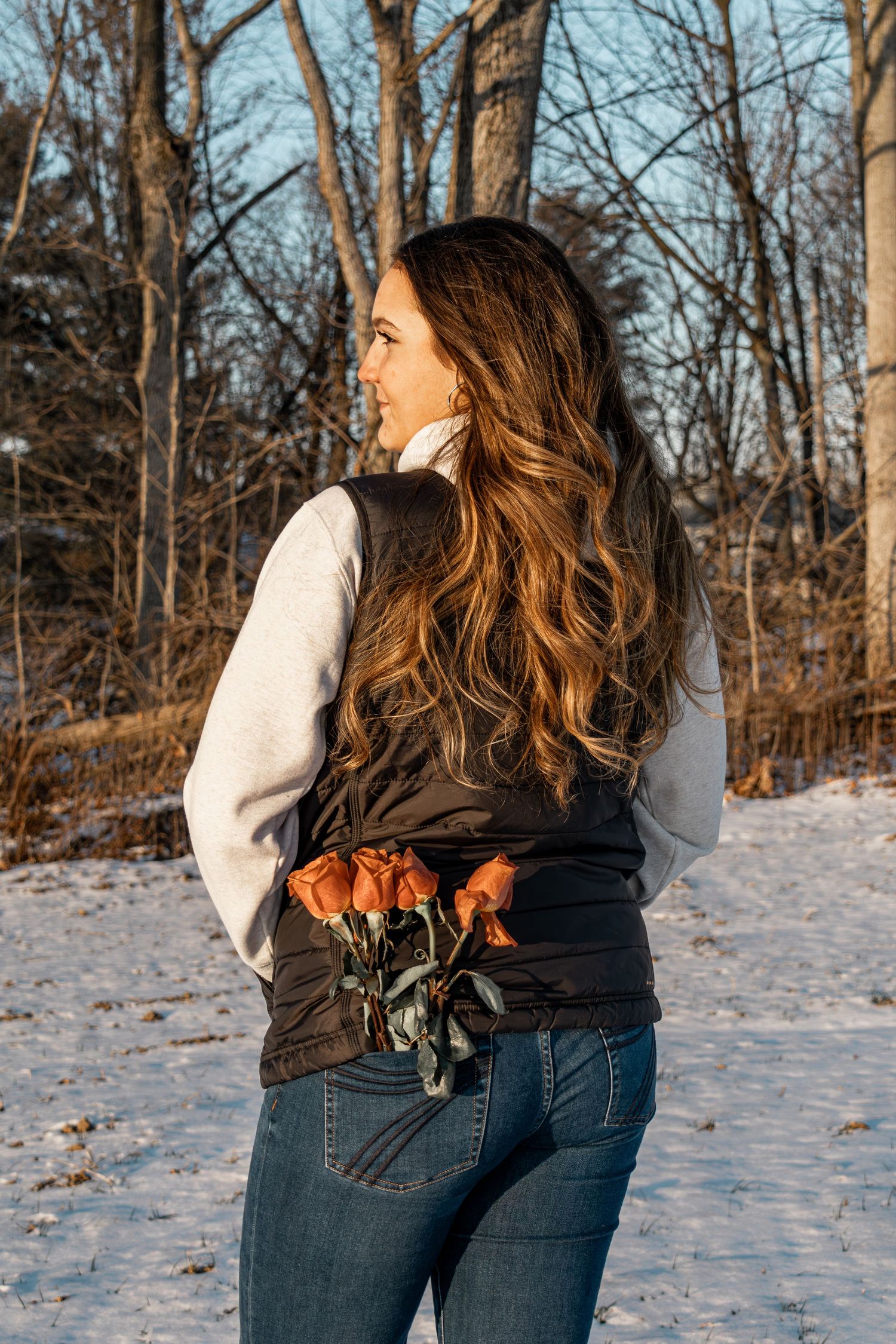 Woman with long wavy hair holding roses behind her back in a snowy winter forest setting.