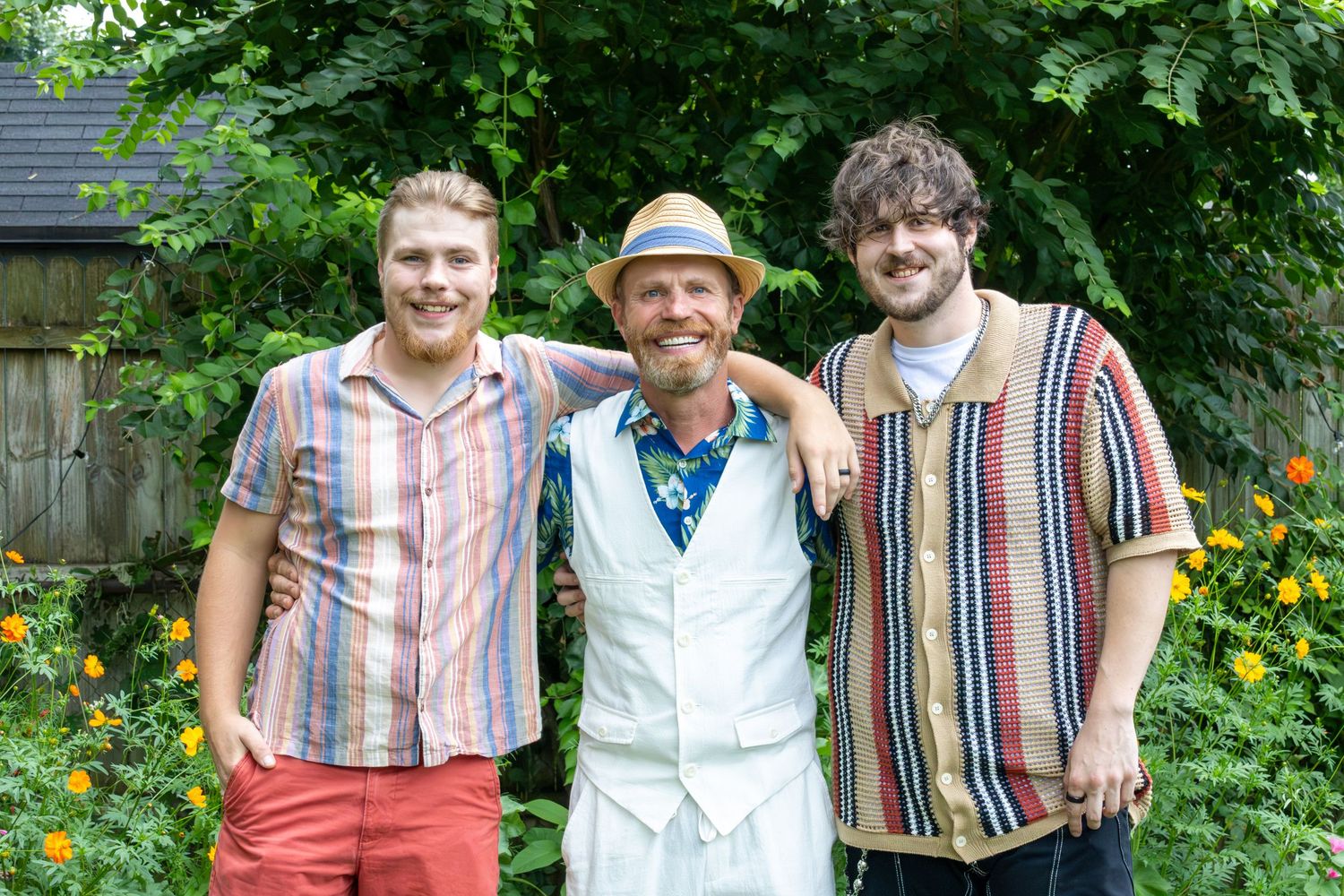 Three smiling men pose together outdoors in a lush green garden, wearing colorful striped and casual summer shirts.
