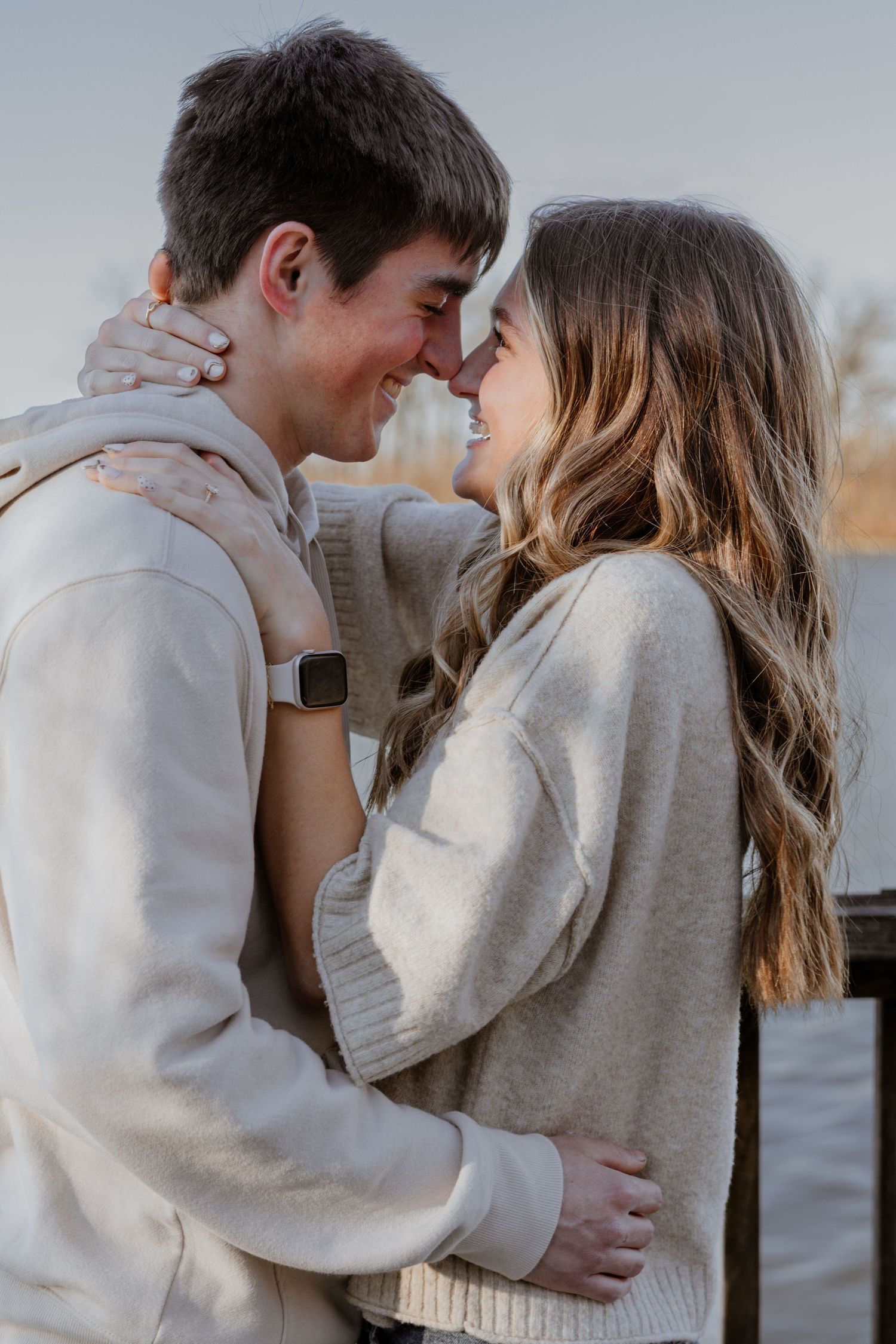 A happy young couple in cream sweaters share a romantic nose-to-nose moment by a waterfront railing.