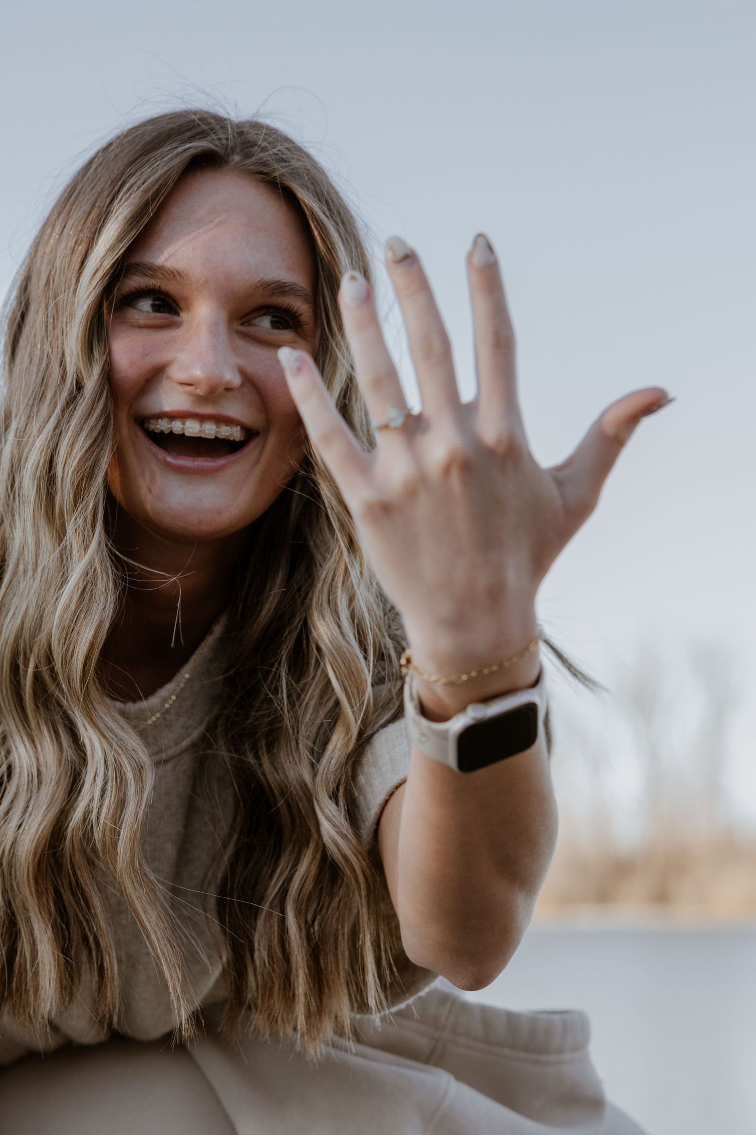Laughing blonde woman showing off engagement ring while wearing a smartwatch outdoors.