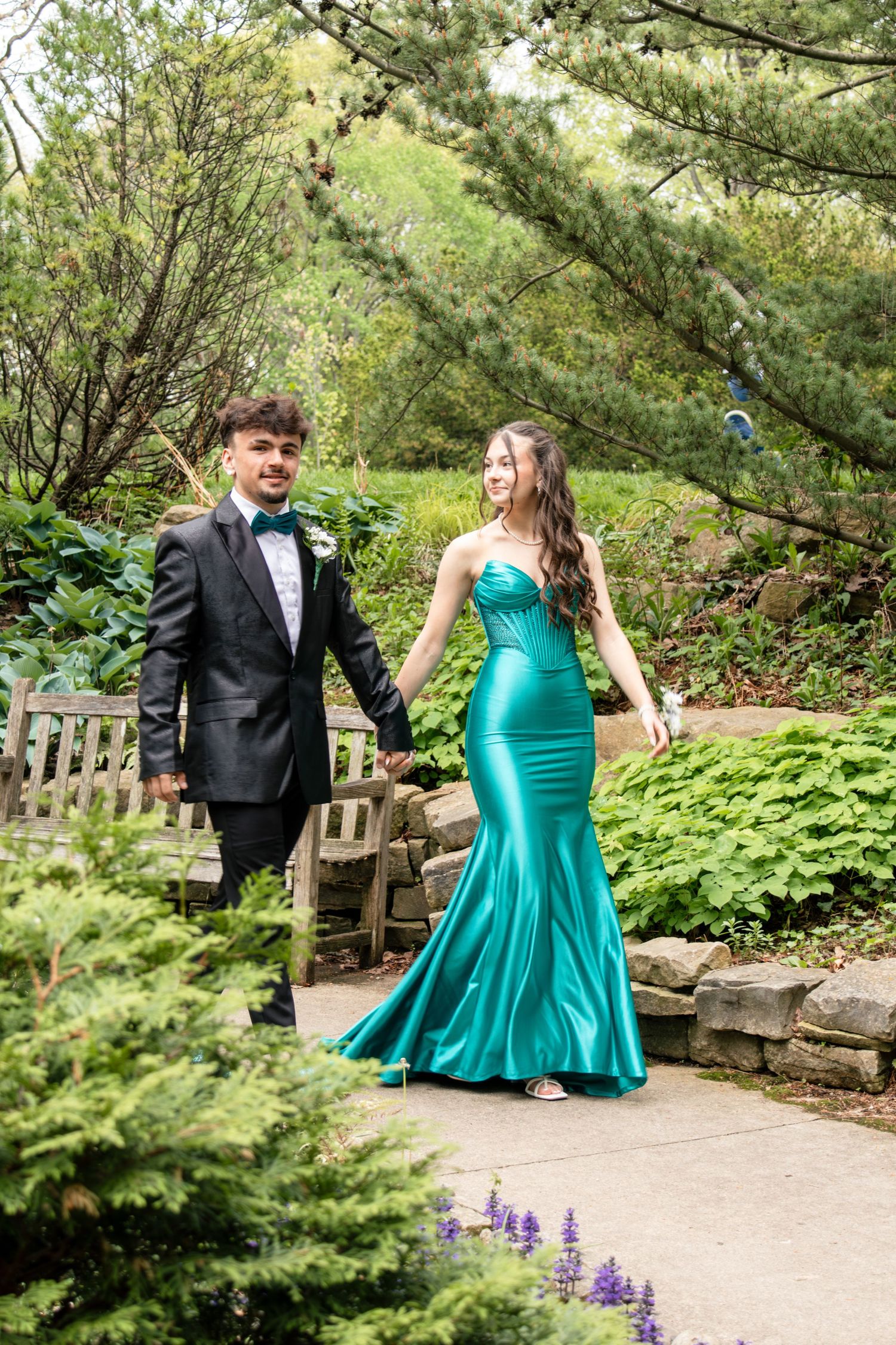 Couple in formal wear holding hands in a lush garden, woman in teal mermaid gown, man in black tuxedo.