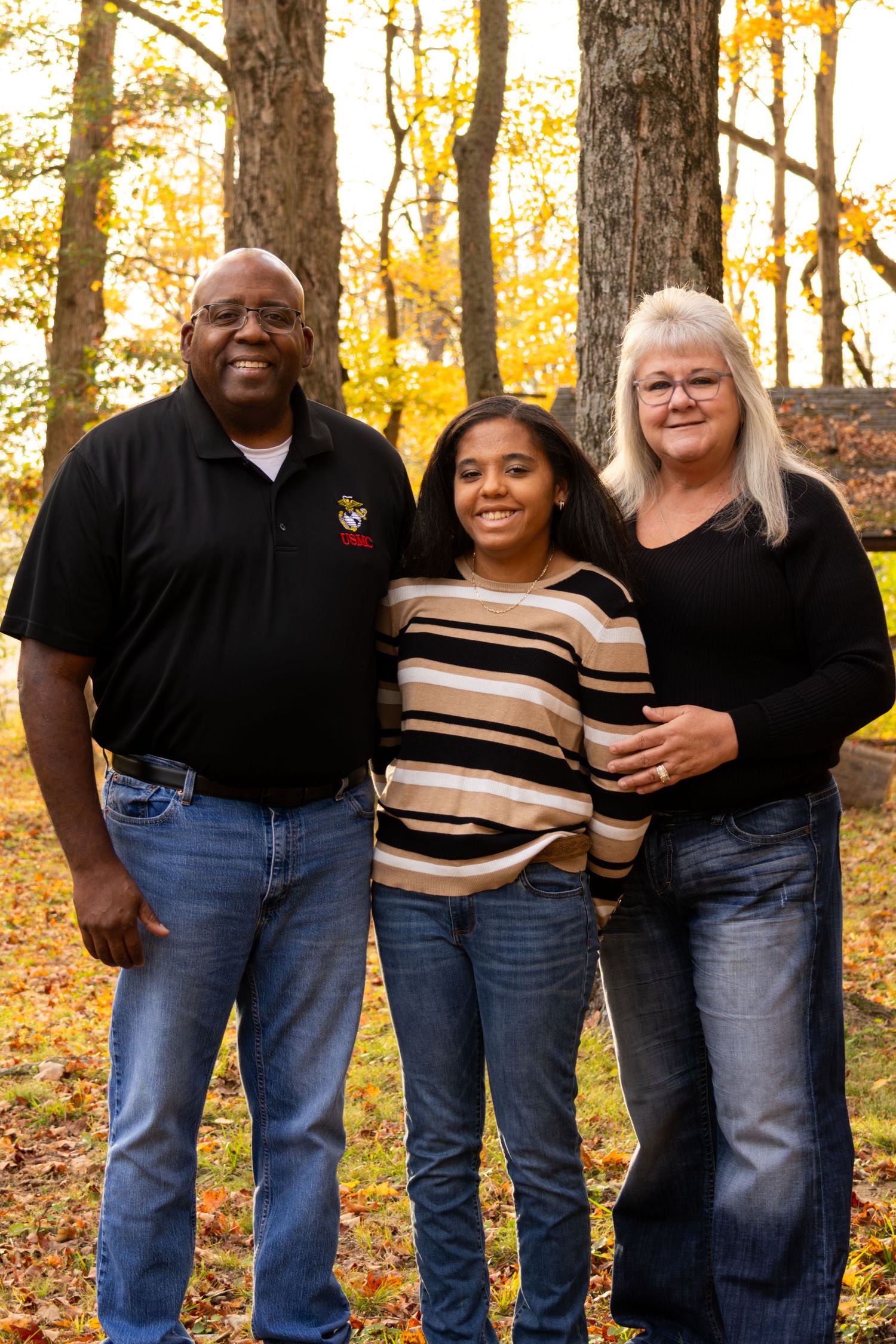 Family of three smiling together in autumn woods with vibrant yellow foliage in the background.