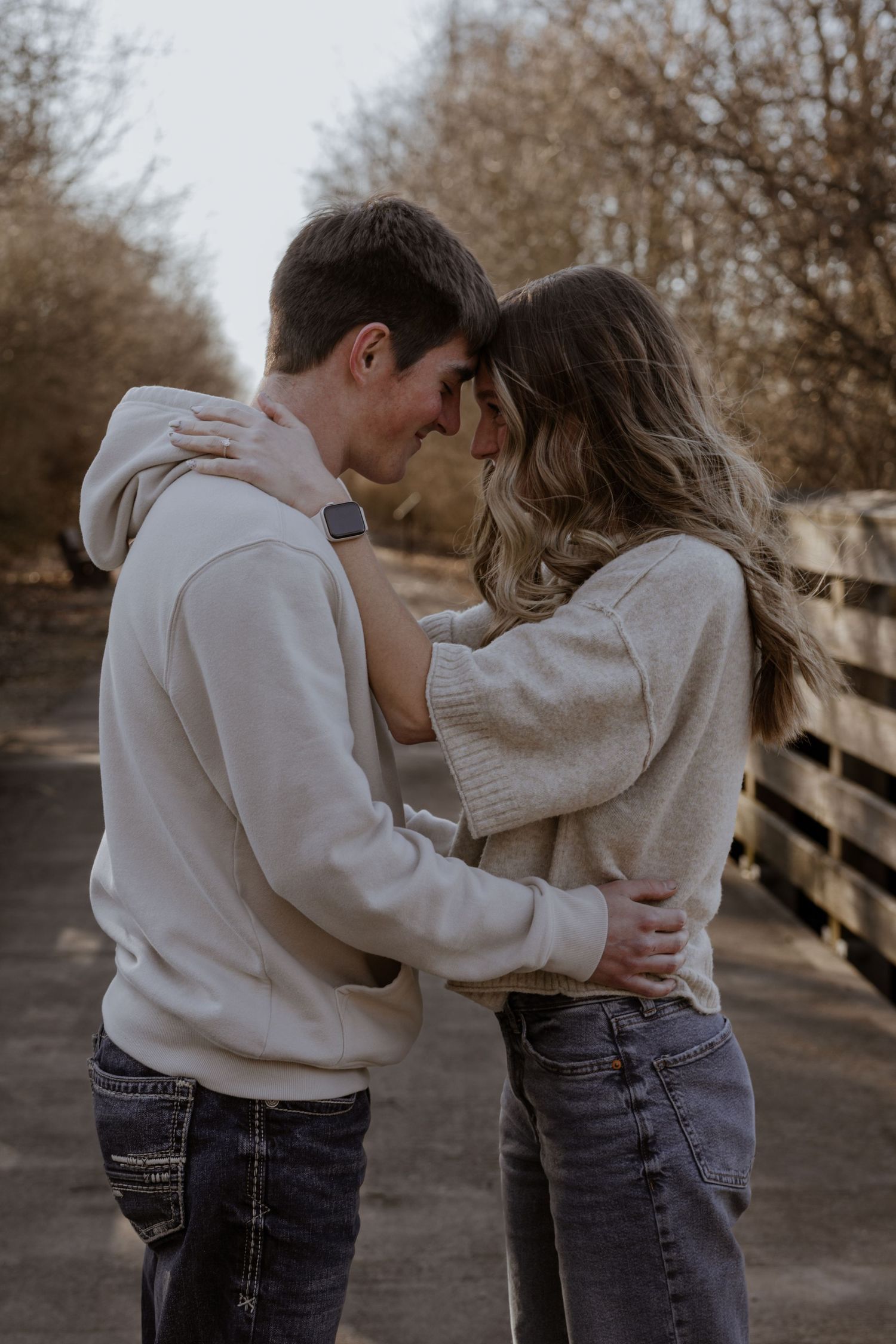 A young couple in matching beige sweaters and jeans embrace tenderly on a rustic wooden bridge path.