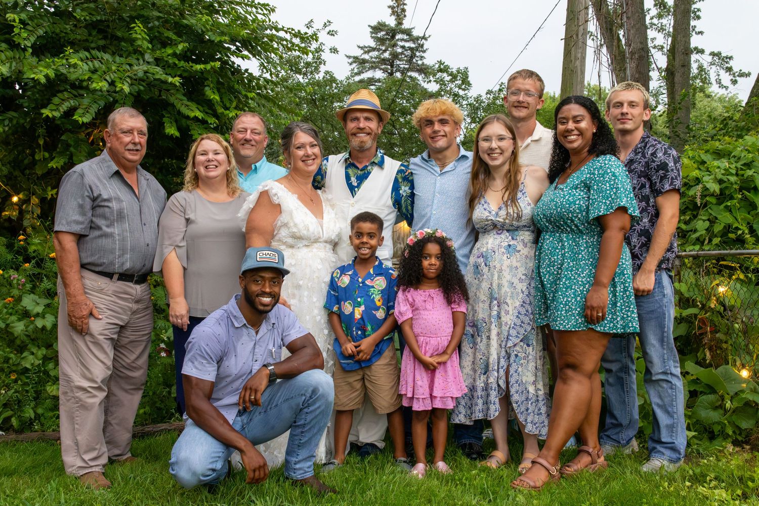 Large diverse family group posing together outdoors at a summer celebration in a lush green backyard.