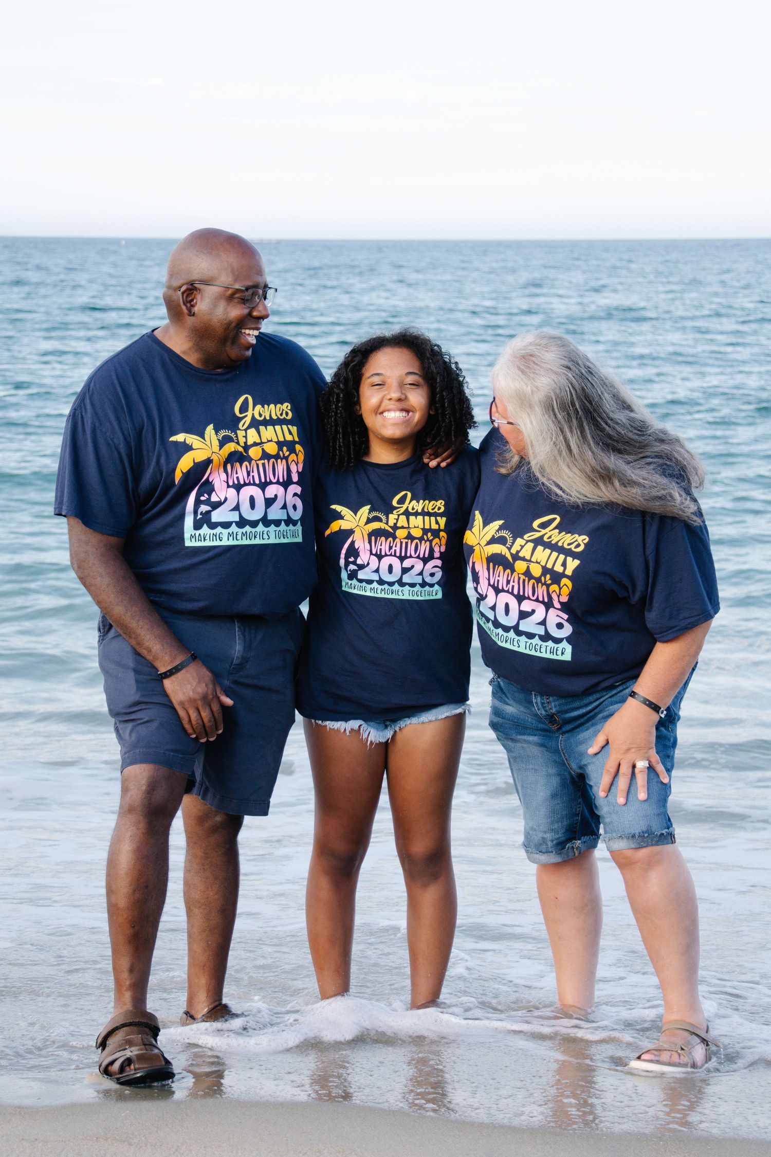 Family of three posing on beach in matching navy Jones Family Vacation 2026 shirts, smiling together at the water's edge.
