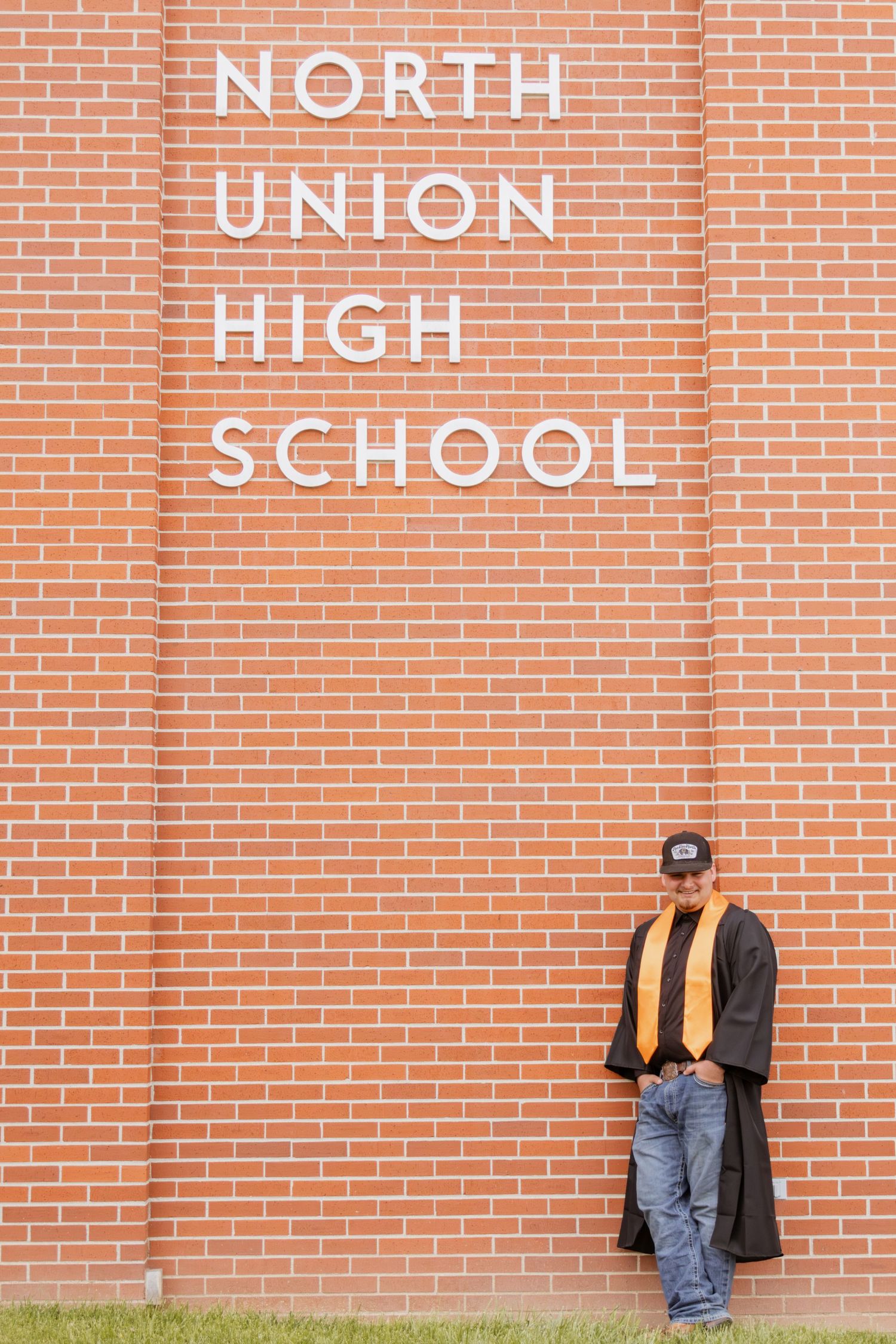 High school graduate in black and gold cap and gown leaning against North Union High School brick wall.