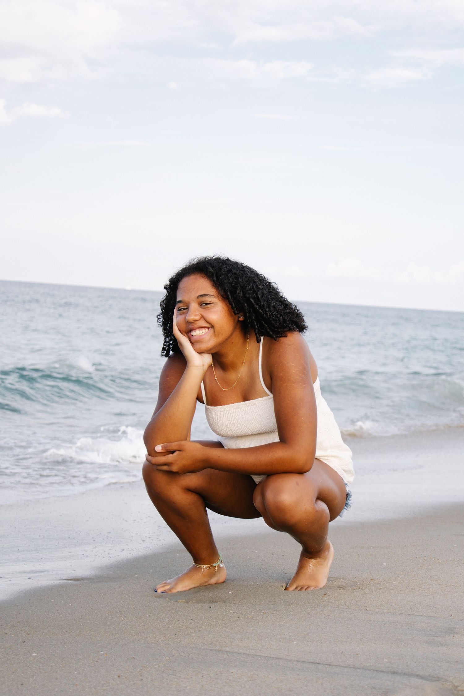 Smiling young woman crouching on a sandy beach with ocean waves in the background.