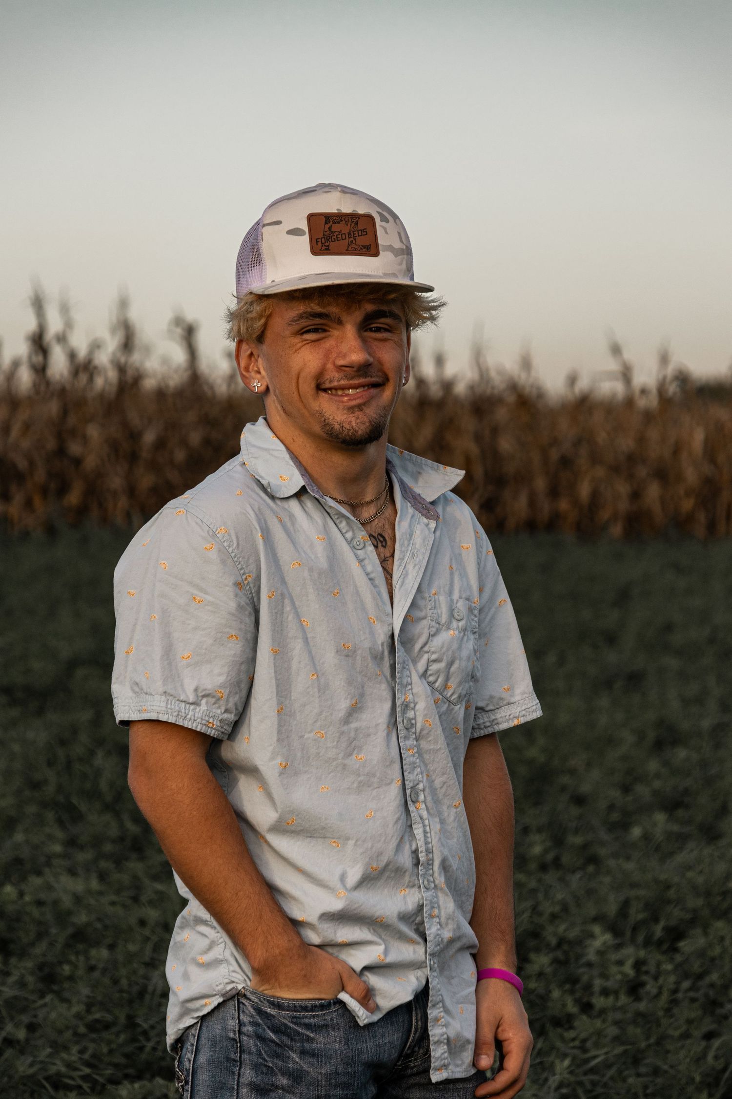 Smiling young man in white cap and light blue shirt standing in farm field at sunset.