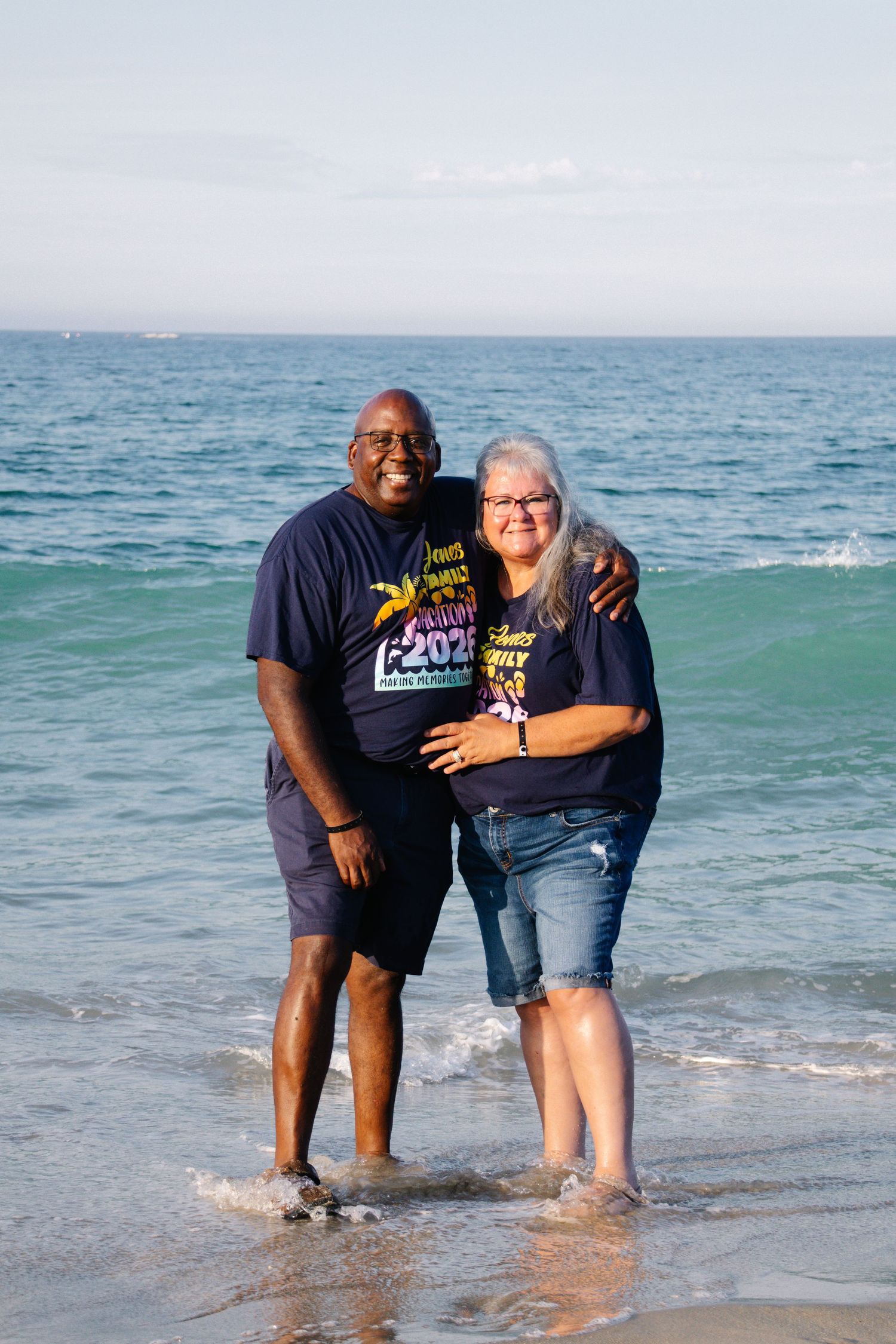 Older couple in matching navy family reunion shirts standing in ocean waves on a beach.