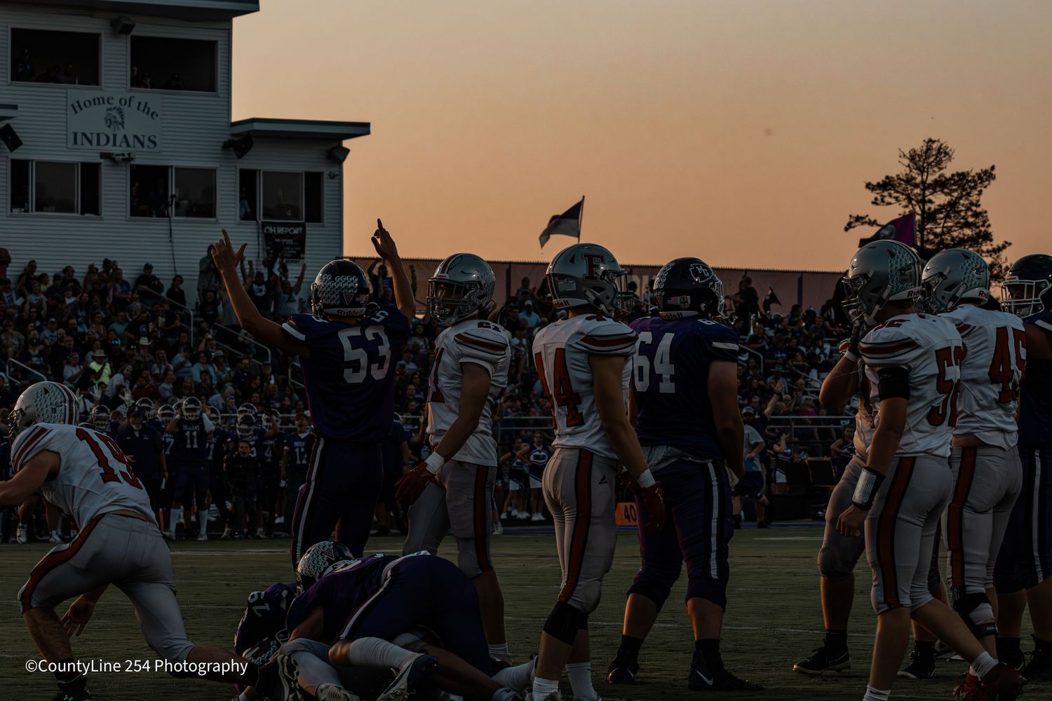 High school football player #53 signals first down during a twilight game with crowd in background.