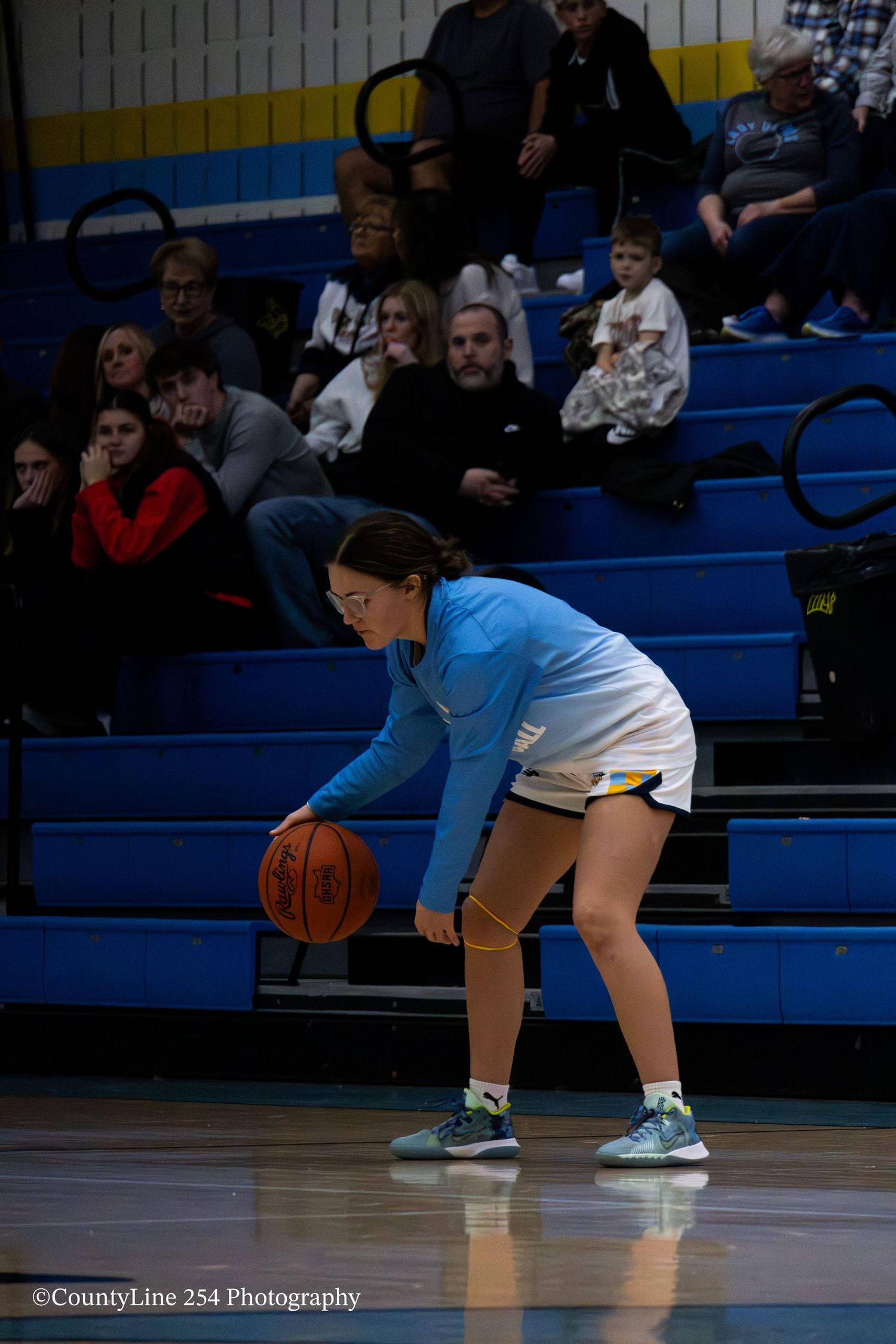 Young female basketball player dribbles ball in defensive stance during a high school gym game.