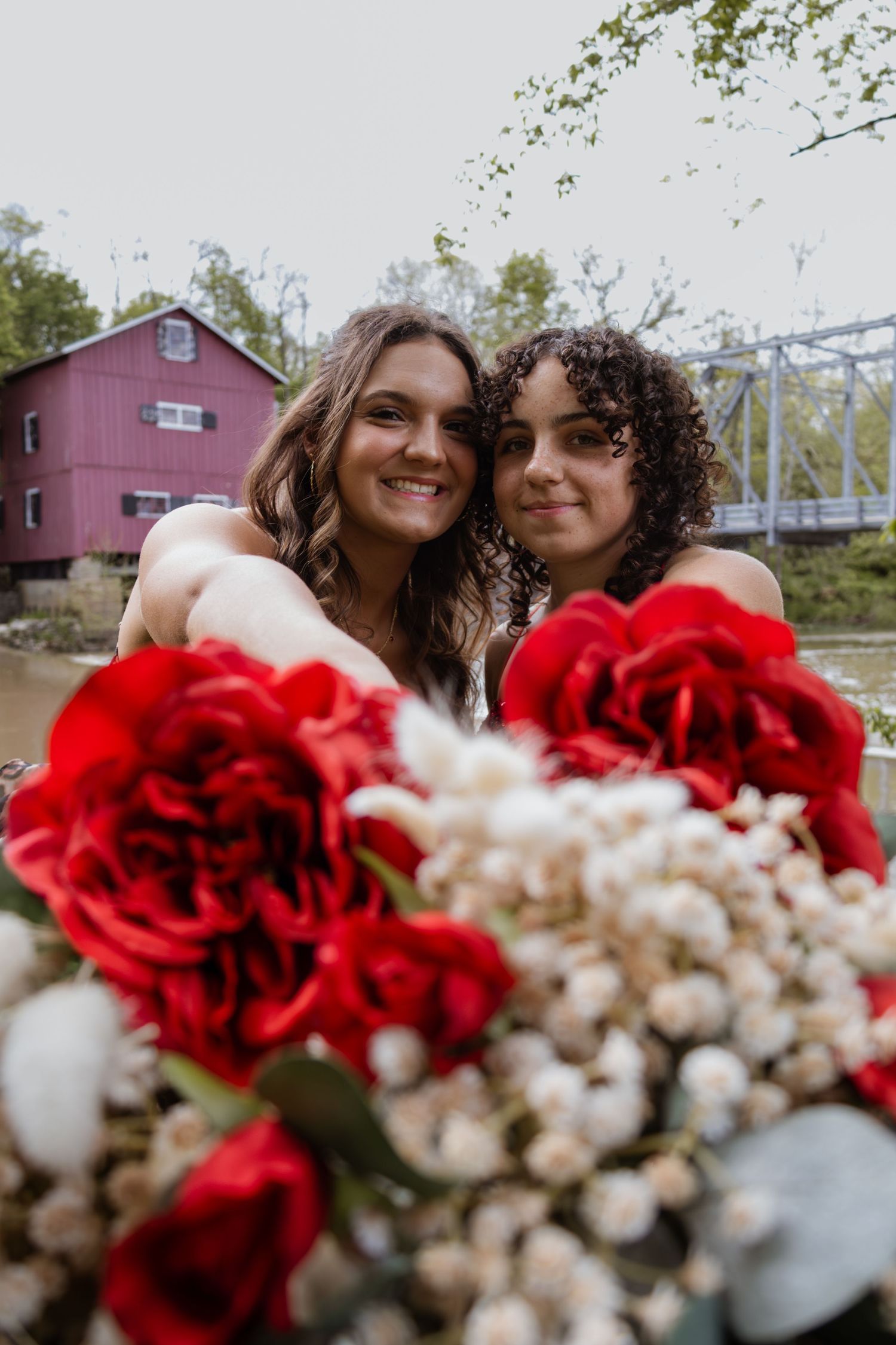 Two smiling young women posing together with red and white floral bouquets near a red barn.