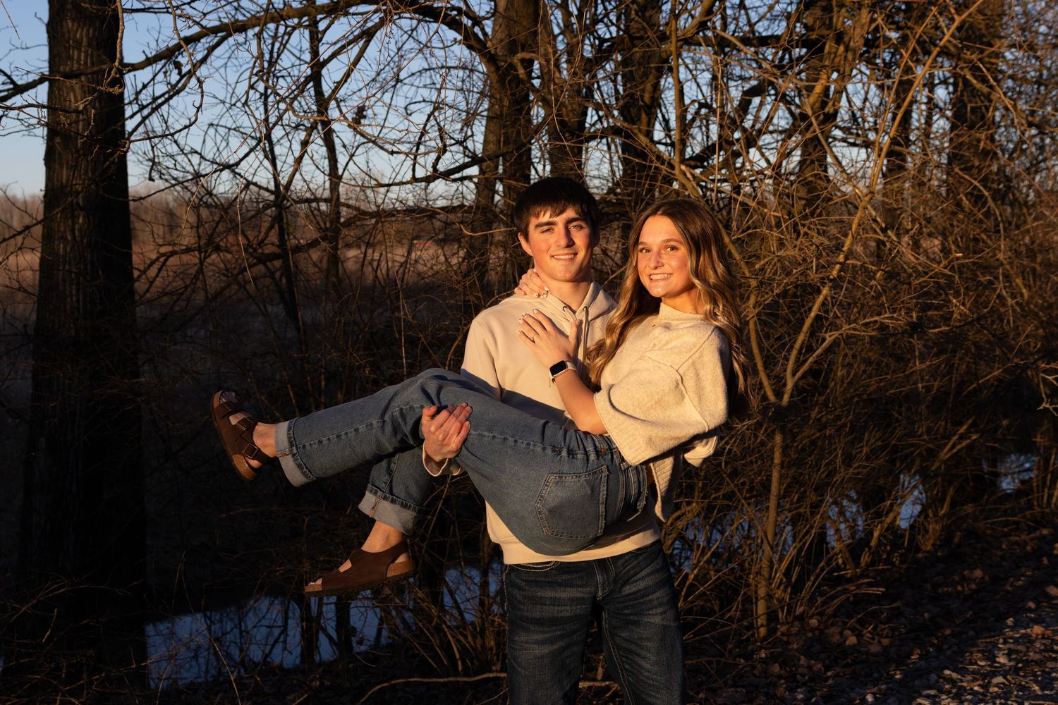 Young couple outdoors at golden hour, man carrying woman in his arms near bare winter trees.