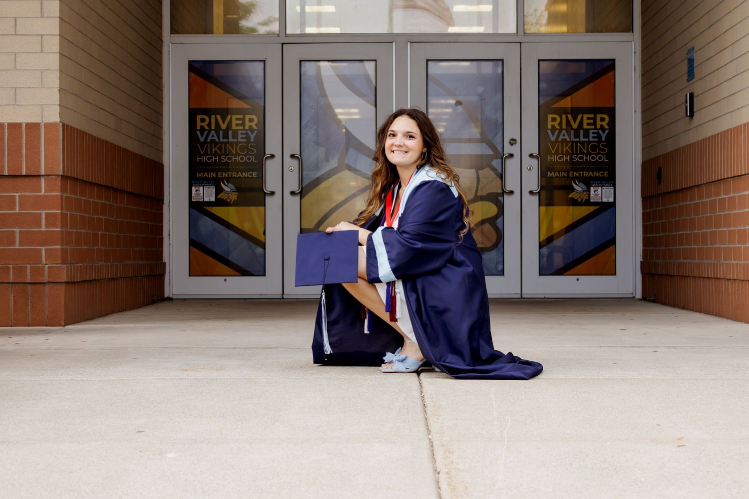 Female graduate in navy cap and gown kneeling outside River Valley Vikings High School entrance.