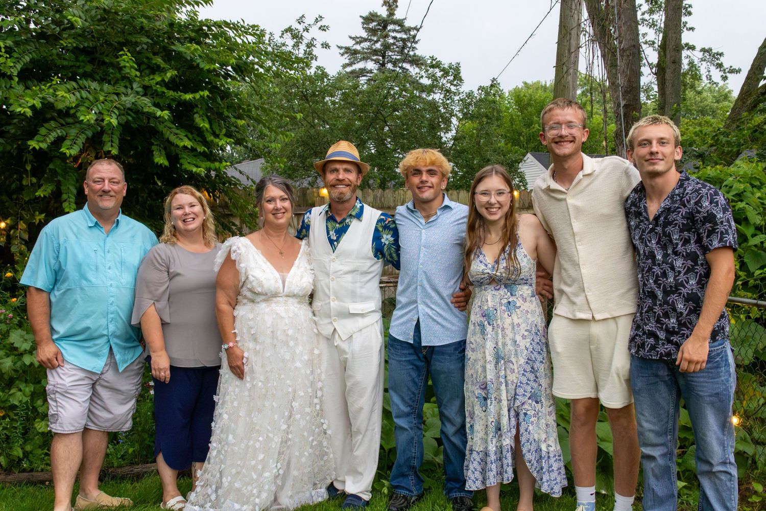 A large group of eight people, including a bride and groom, pose together outdoors at a summer wedding celebration.