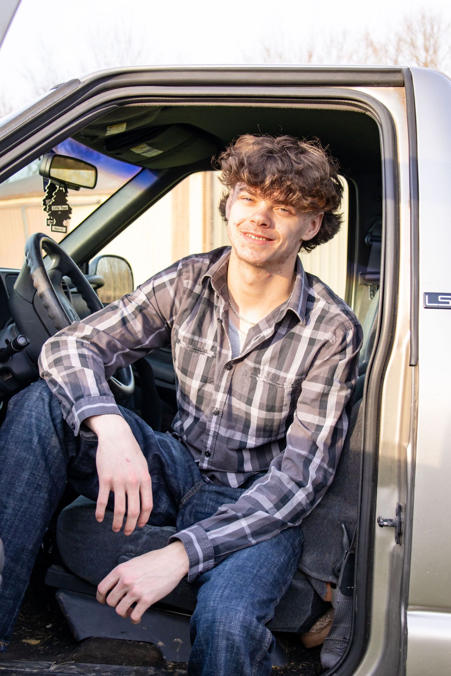 Smiling young man in plaid shirt sitting relaxed in the open door of a pickup truck.