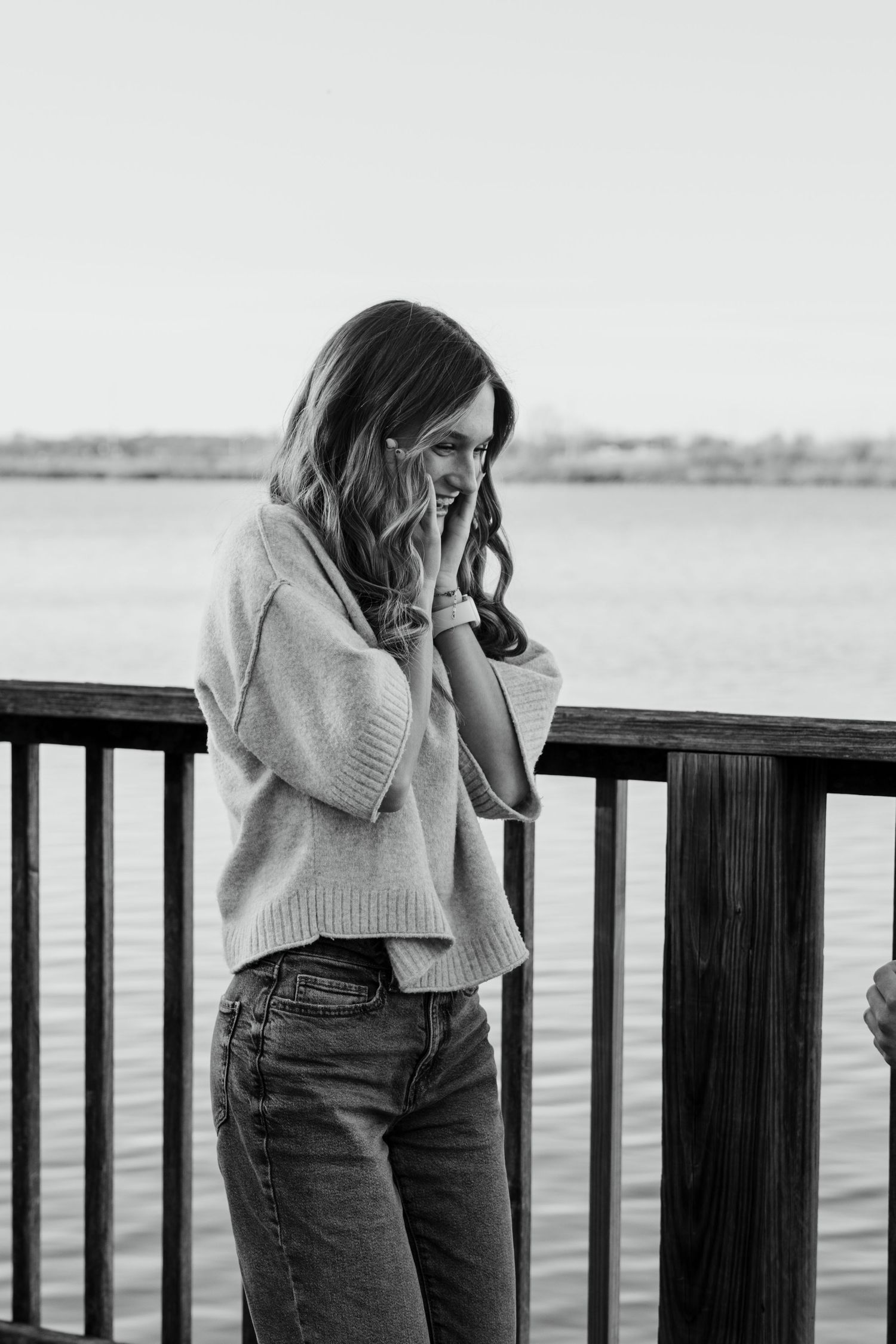 Black and white portrait of a smiling young woman leaning on a railing overlooking water.