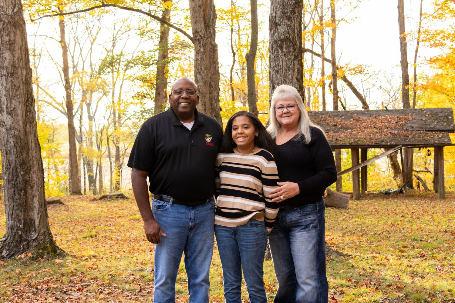 A family of three poses warmly together in a fall park setting with golden autumn leaves and trees in the background.