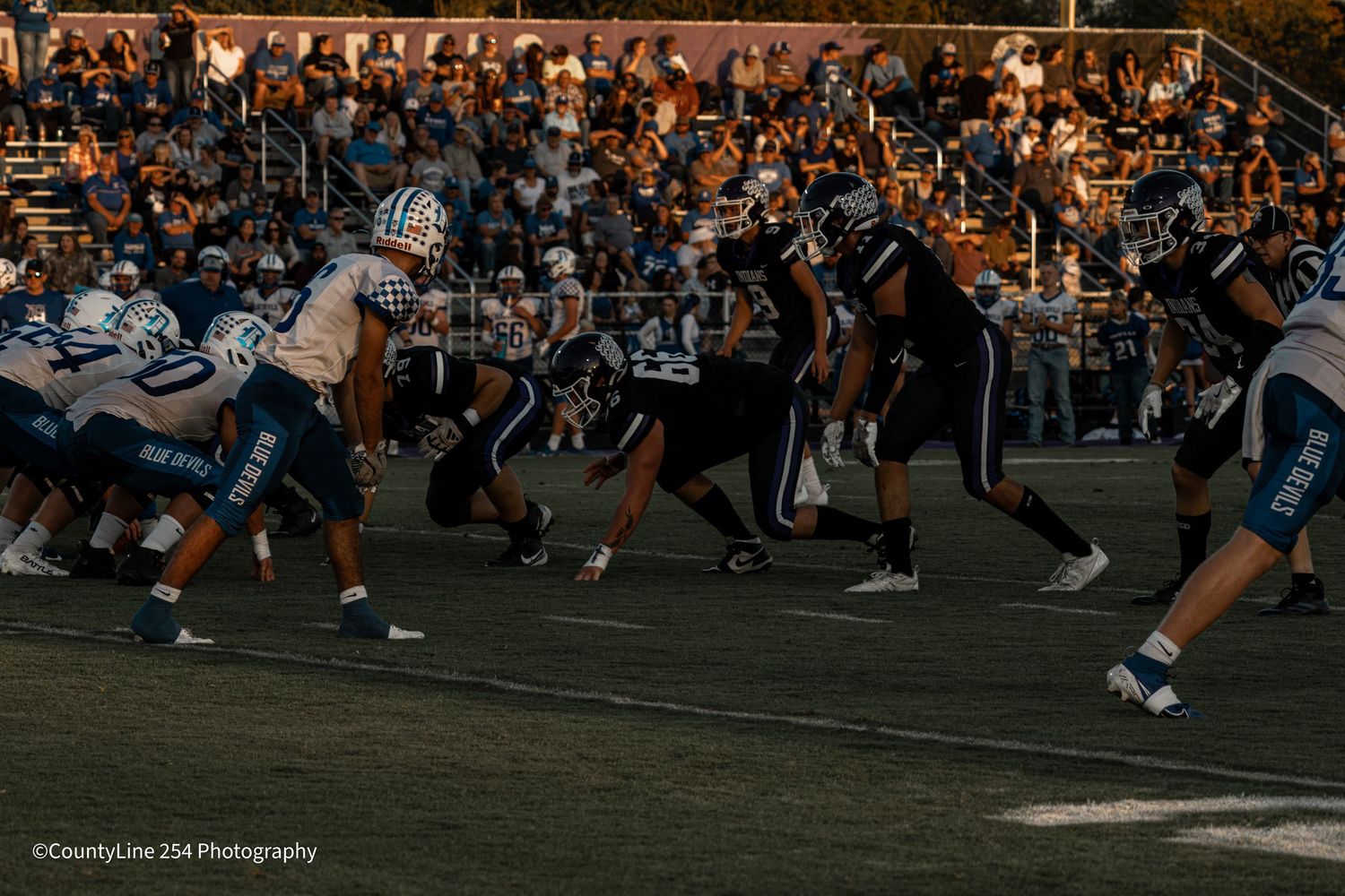 Two football teams line up at the line of scrimmage under bright stadium lights before a snap.