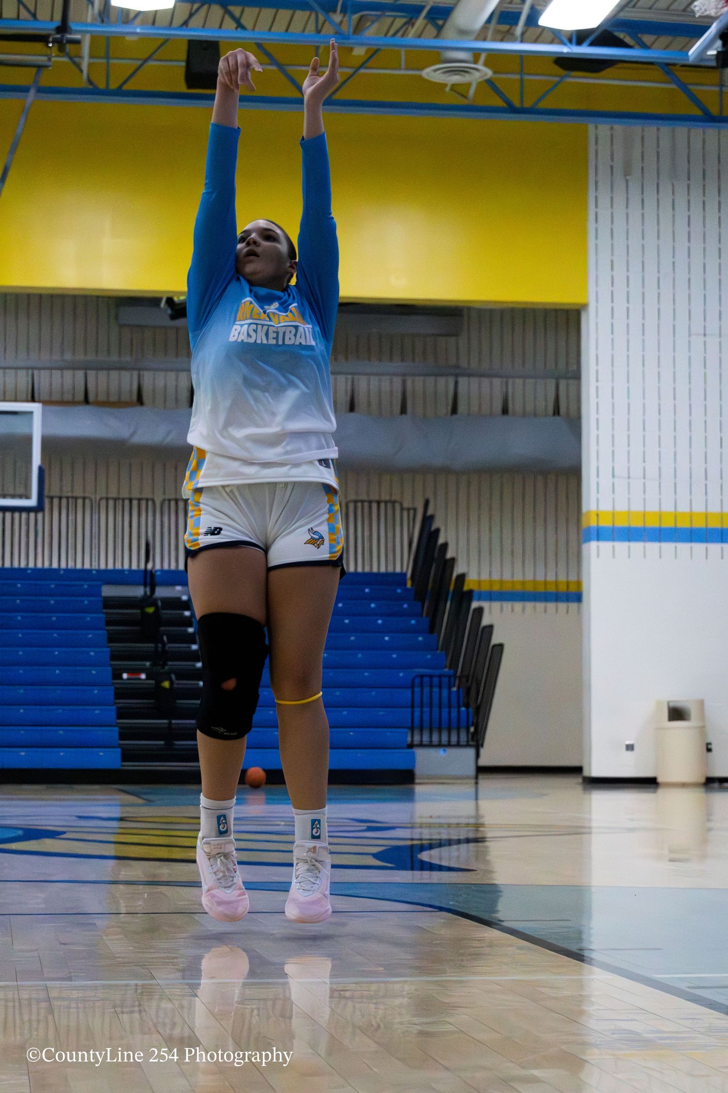 A female basketball player in blue uniform shoots a jump shot during warm-ups in a gymnasium.