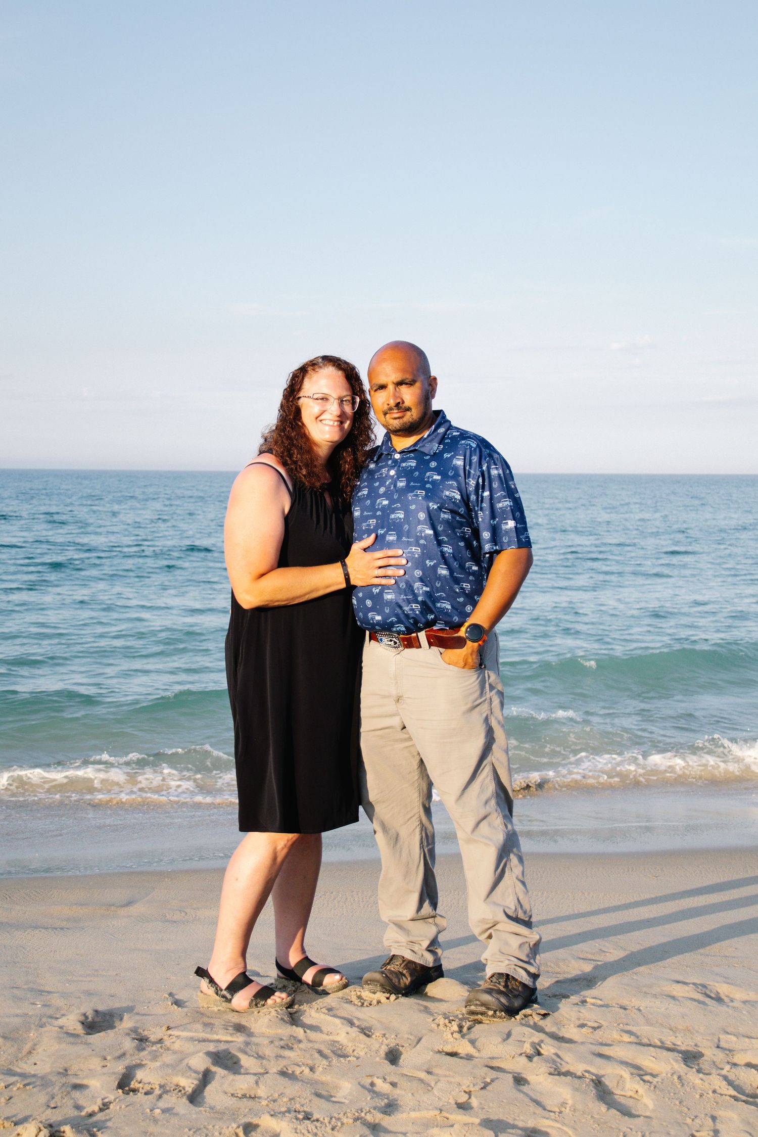 Couple posing together on a sandy beach at sunset, woman in black dress and man in blue floral shirt.
