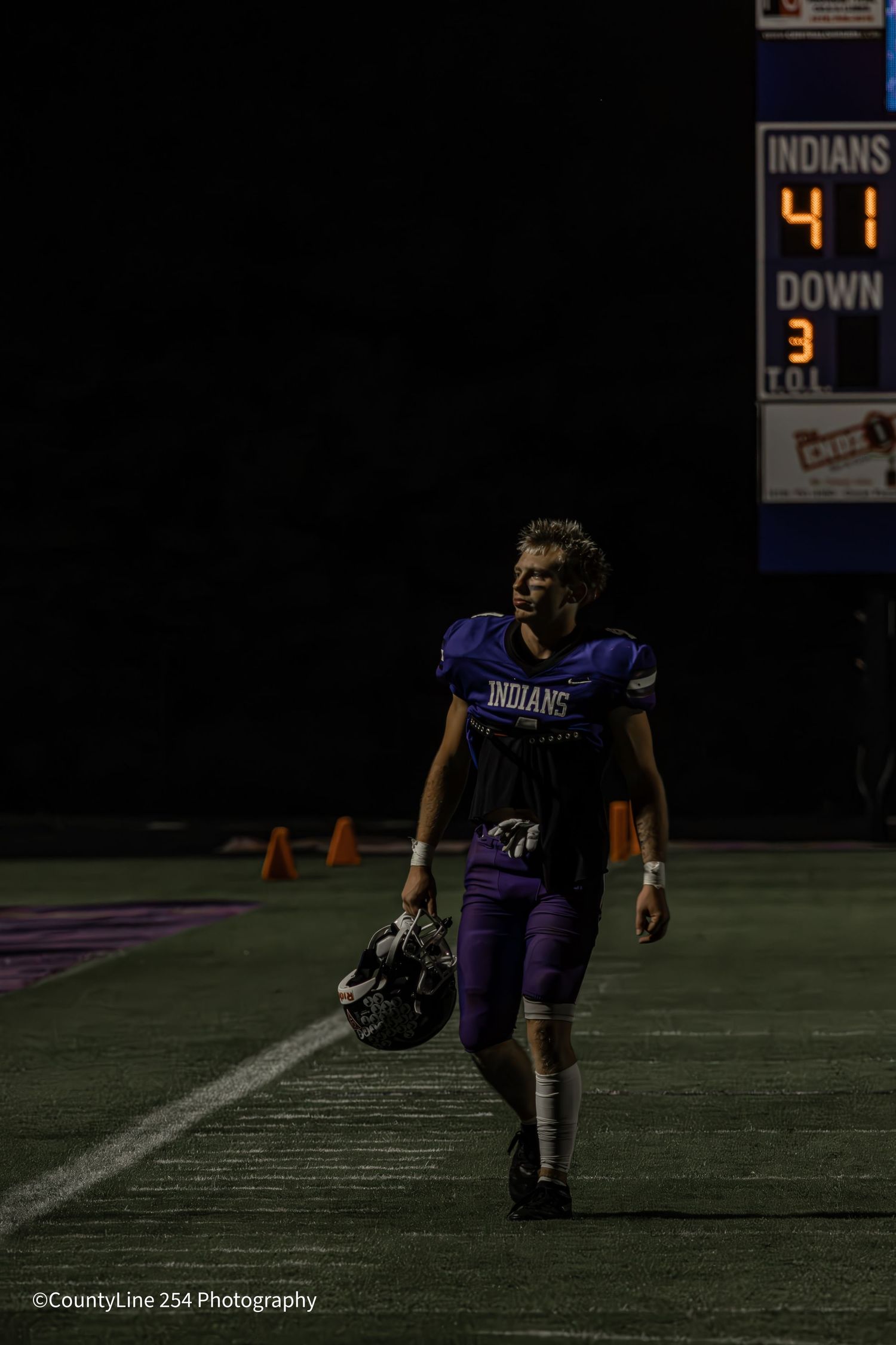 High school football player in purple Indians uniform walks alone on field holding helmet at night.