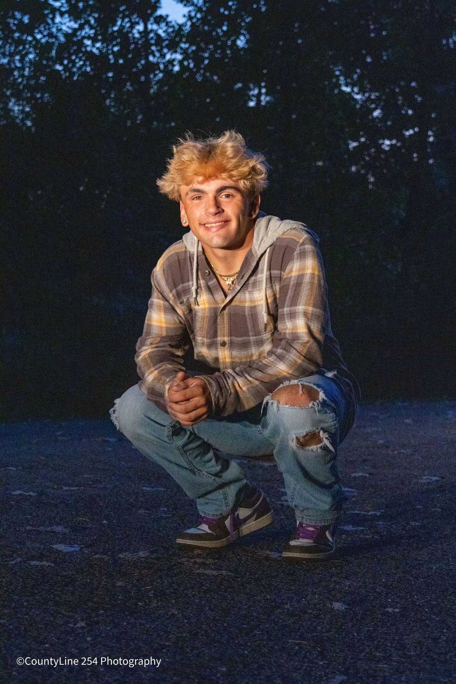 Blonde teen boy crouching in ripped jeans and plaid hoodie on pavement at dusk.