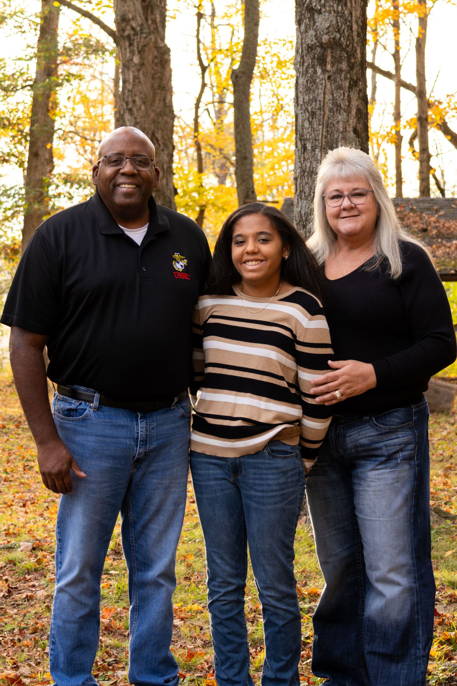 Family of three smiling together outdoors among colorful autumn foliage, wearing casual black tops and jeans.