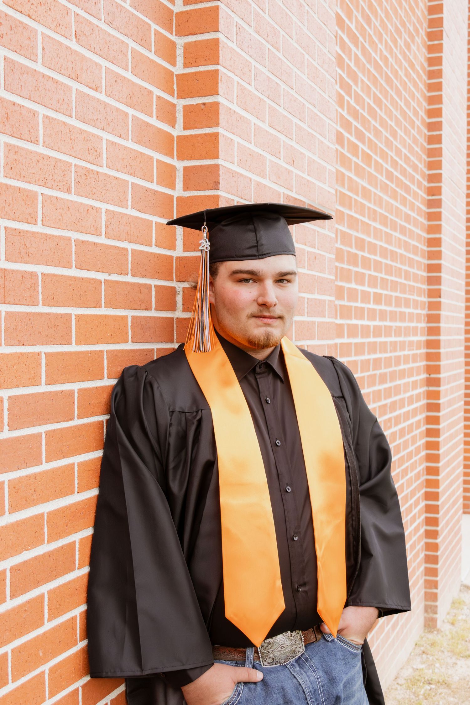 Young male graduate in black cap and gown with gold stole posing against a red brick wall.