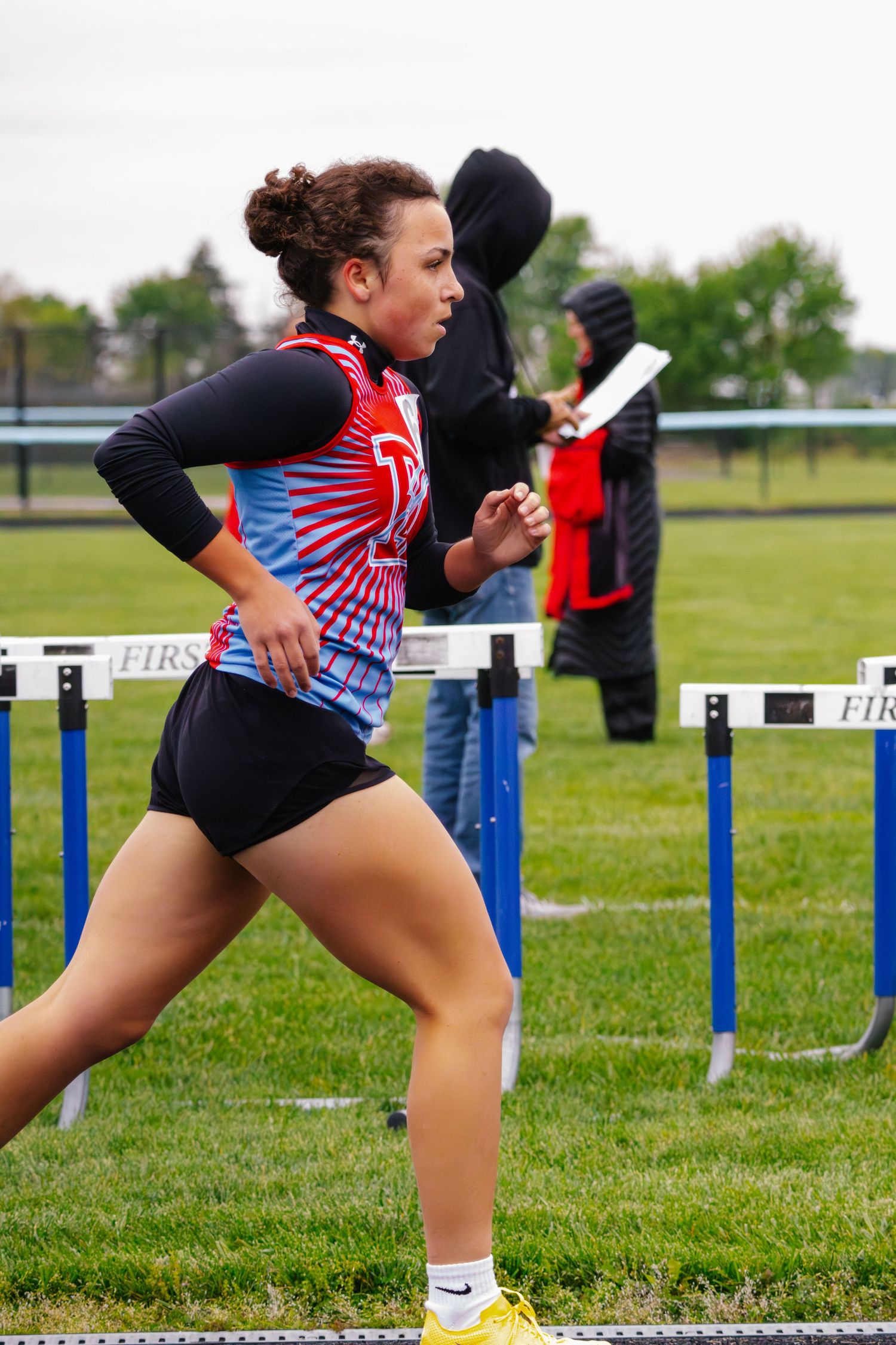 Female track athlete in red and blue uniform running hurdles at outdoor track meet.