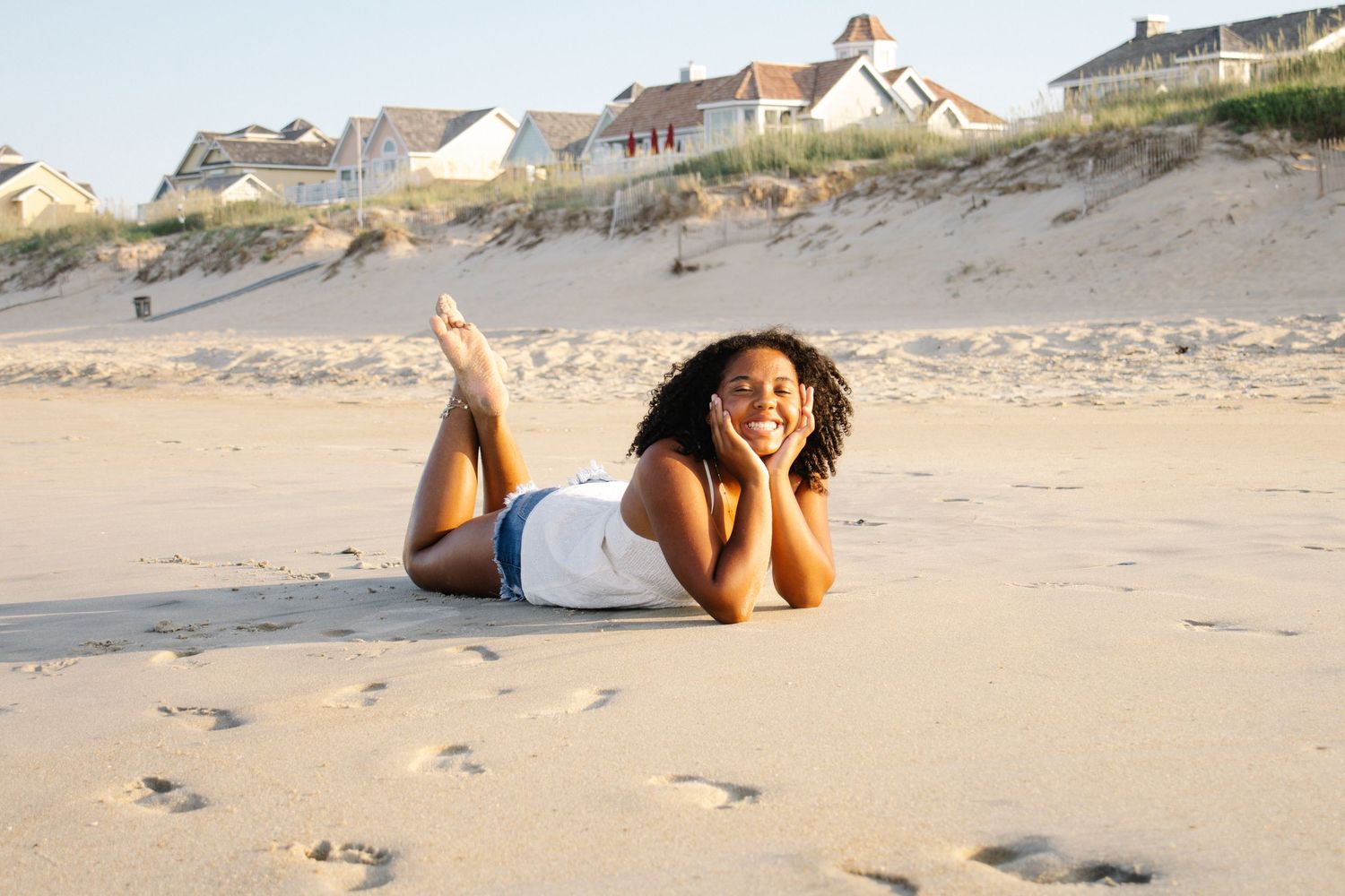 A young Black woman in a white top relaxes on a sandy beach, smiling in golden hour sunlight.