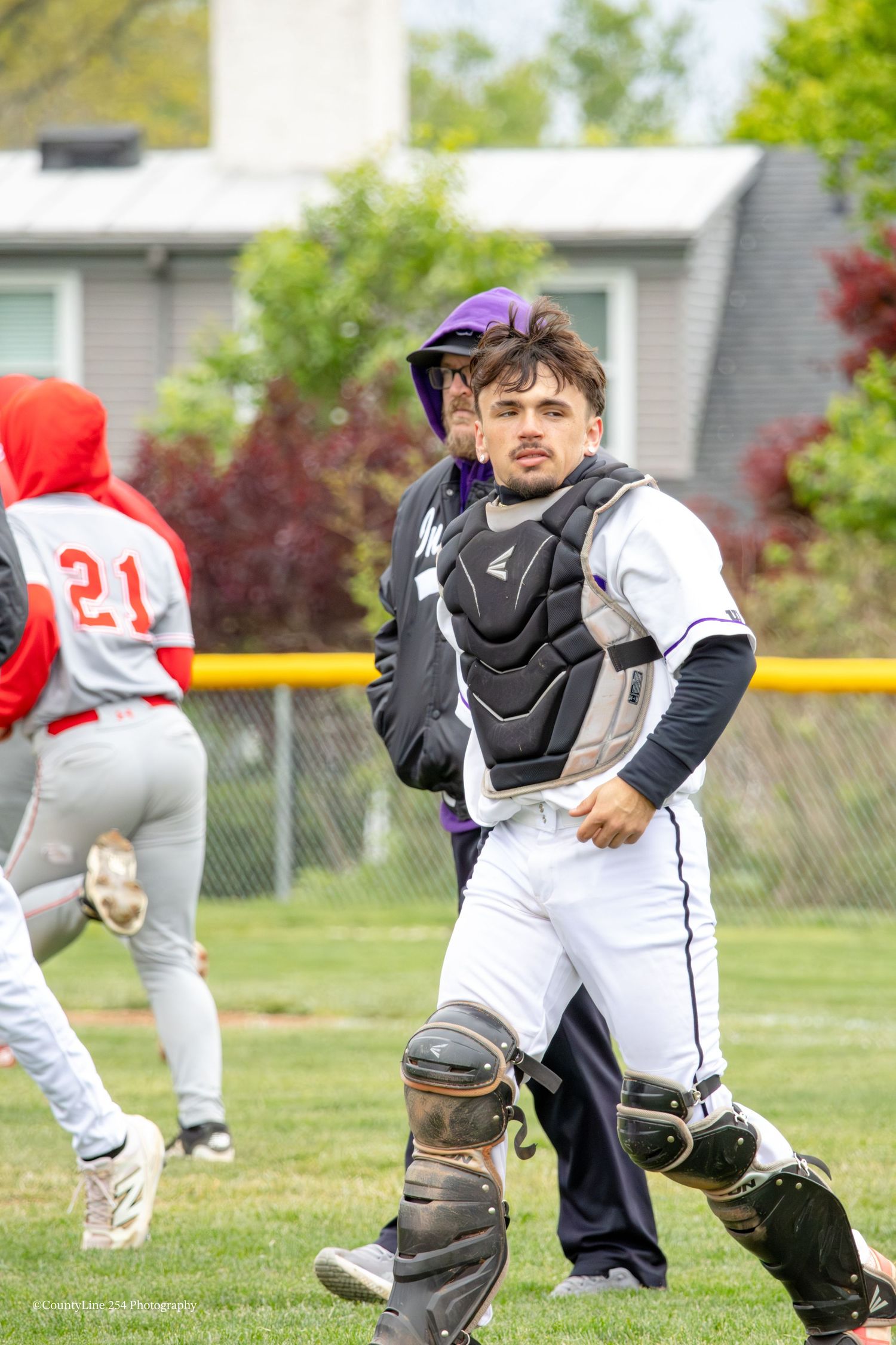 High school baseball catcher in full protective gear watches play while coach stands behind him.