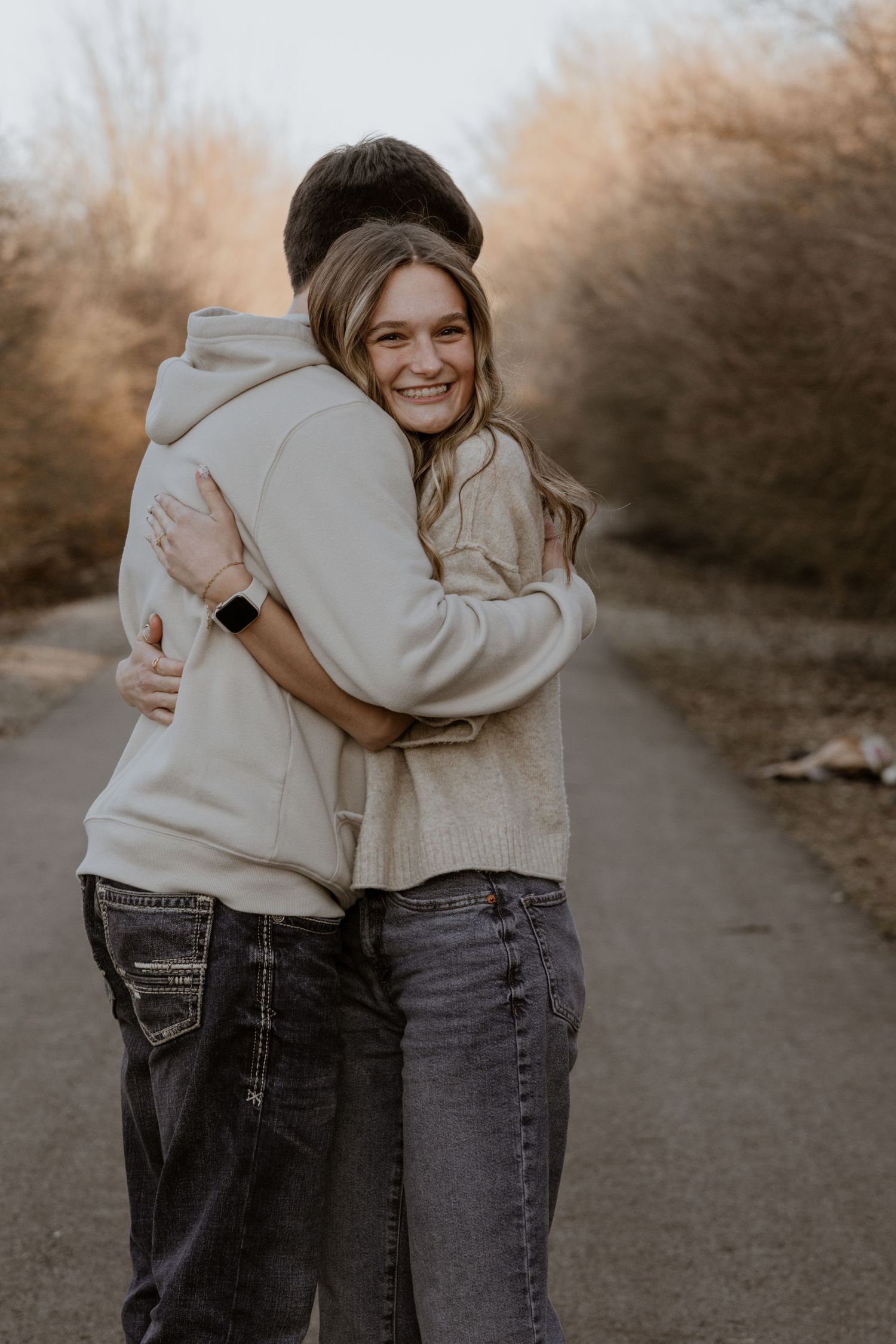 A smiling young woman in a cream sweater hugs her partner on a tree-lined path in warm sunlight.