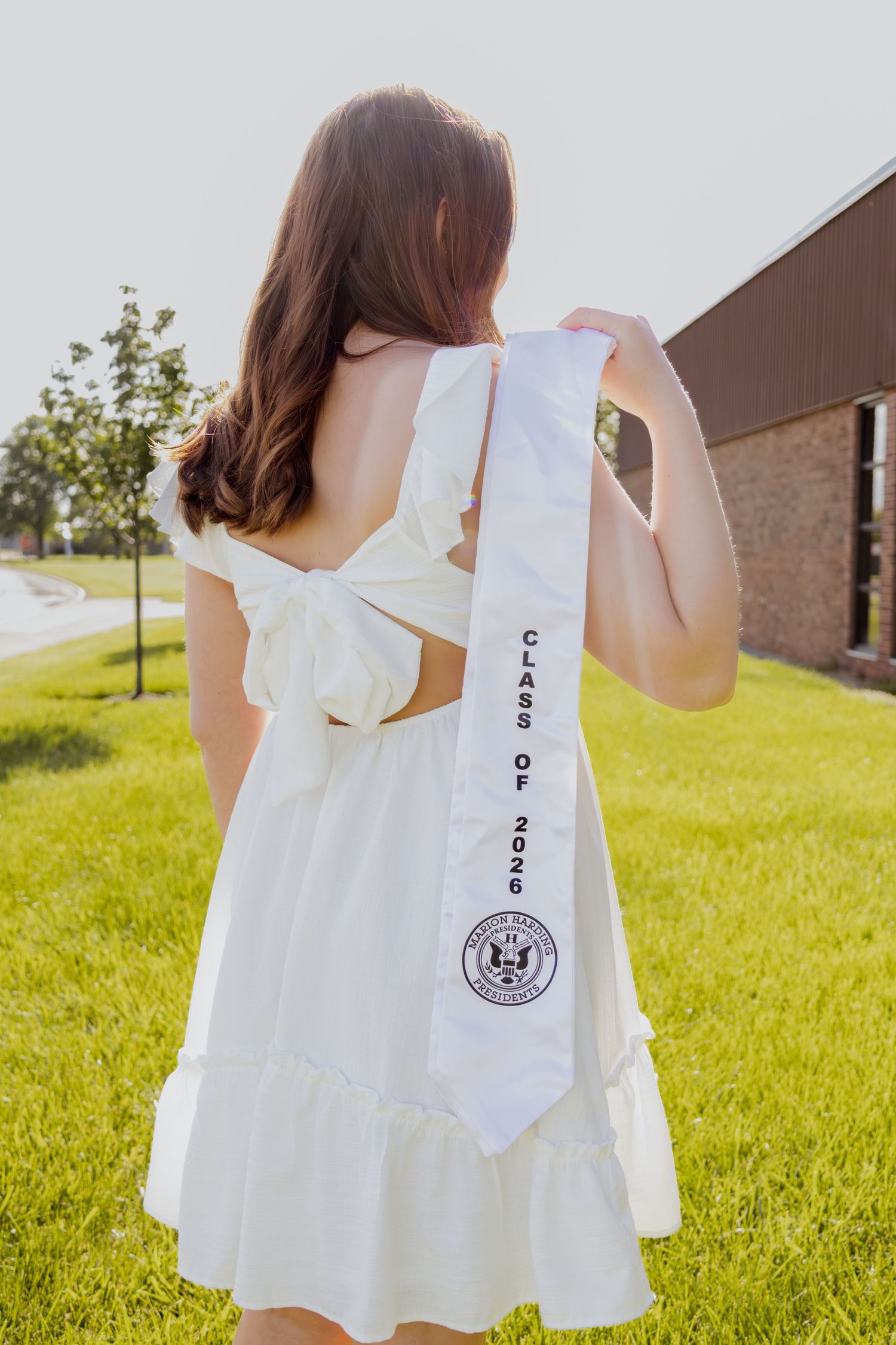 Senior girl in white dress displaying Class of 2026 graduation sash from behind outdoors.