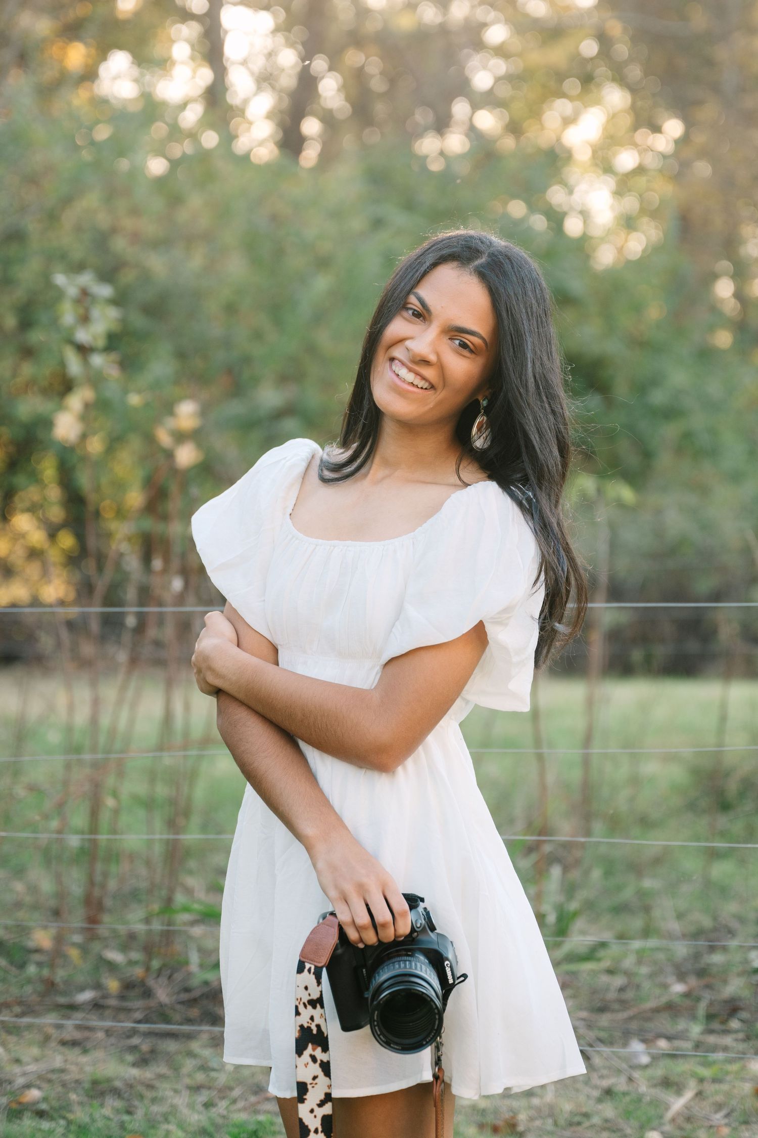 Smiling young woman in white dress holding a DSLR camera outdoors in golden hour light.