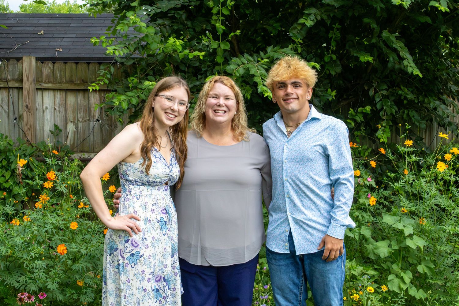 A woman and two teenagers smile together in a lush backyard garden filled with bright yellow flowers in summer.