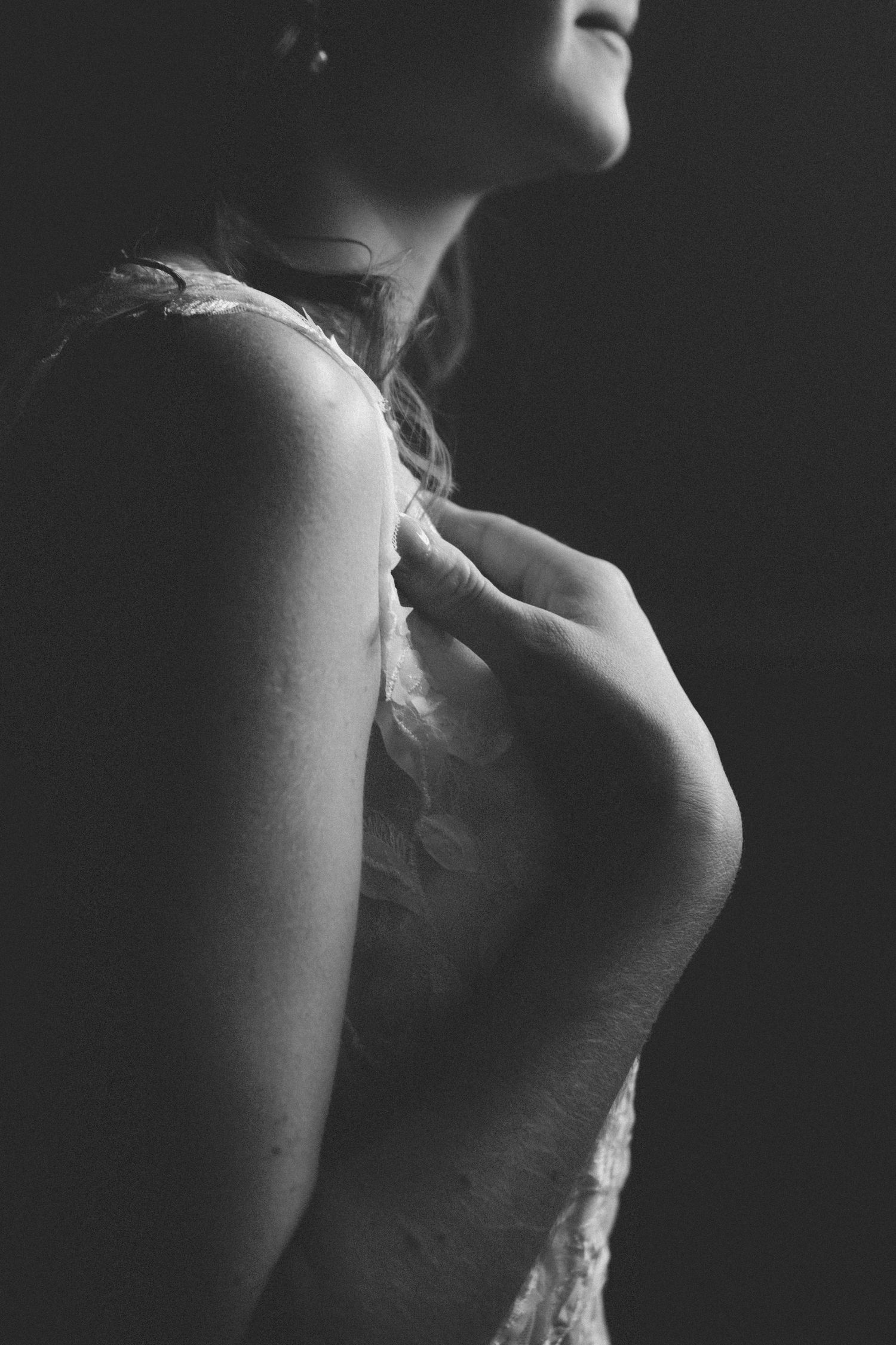 Black and white photo of a woman in a lace dress adjusting her shoulder strap against a dark background.