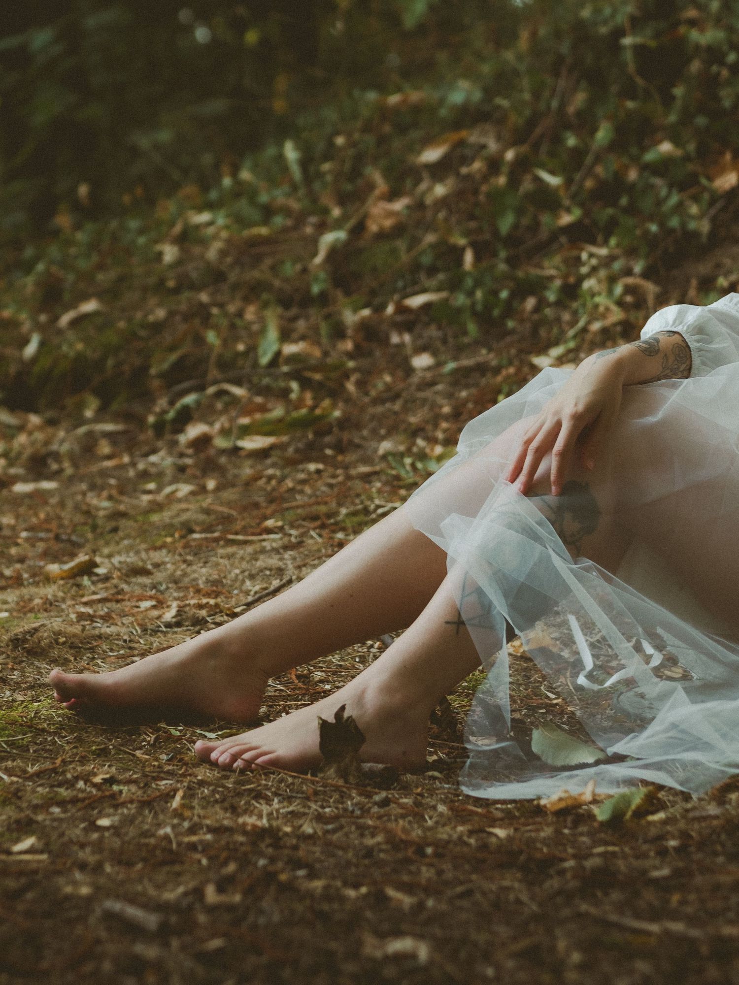 Barefoot woman in white tulle dress sitting on forest ground among fallen leaves.