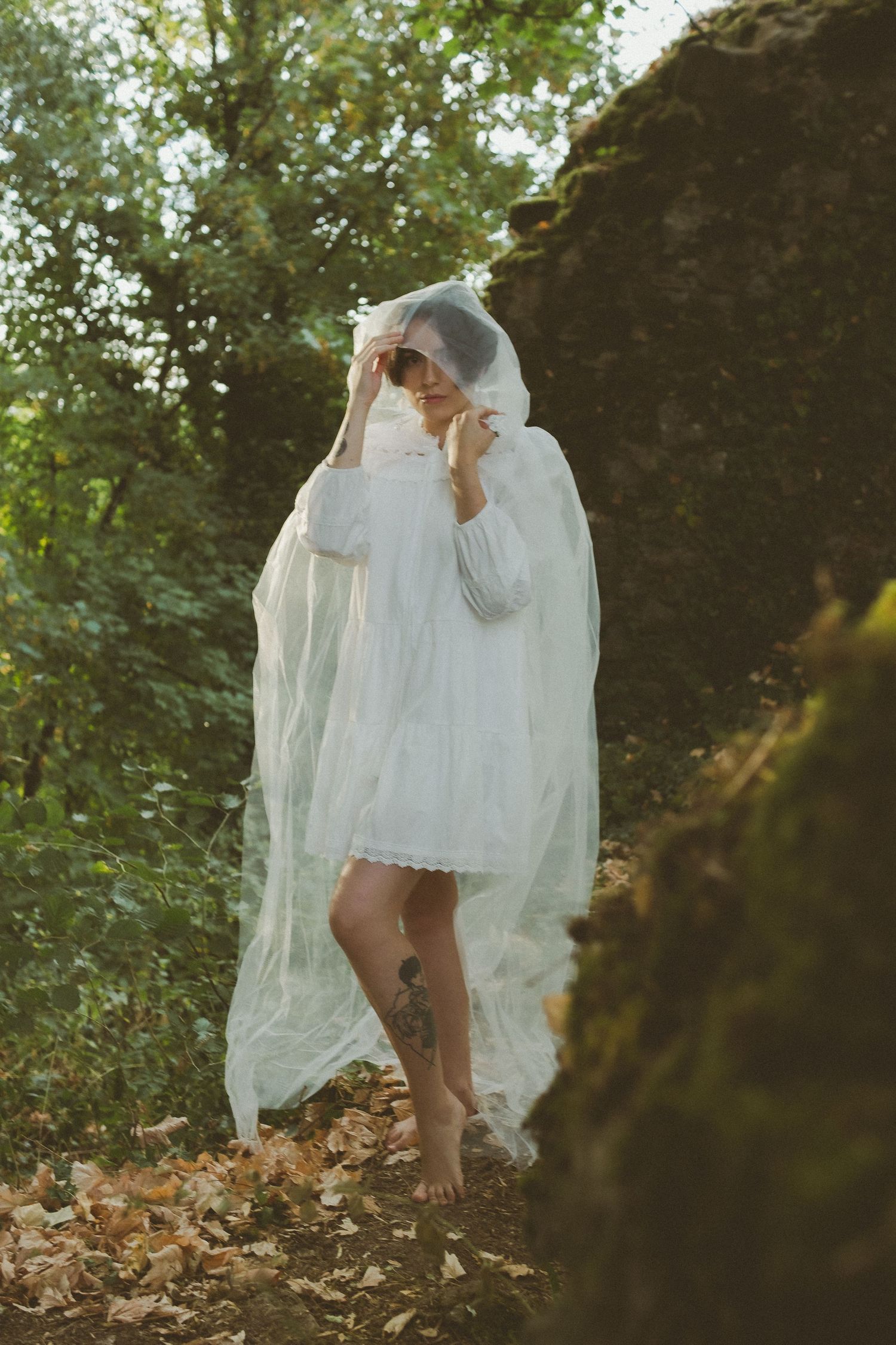 Barefoot woman in white dress draped with sheer veil stands among mossy forest rocks.