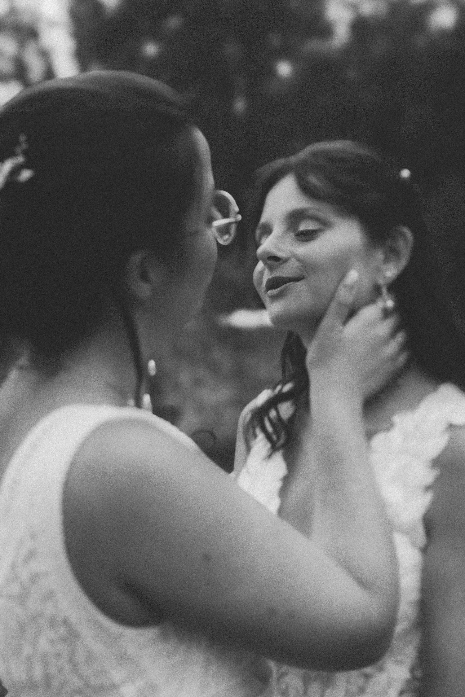 Black and white photo of two brides in white lace dresses sharing a tender moment, one gently cupping the others face.