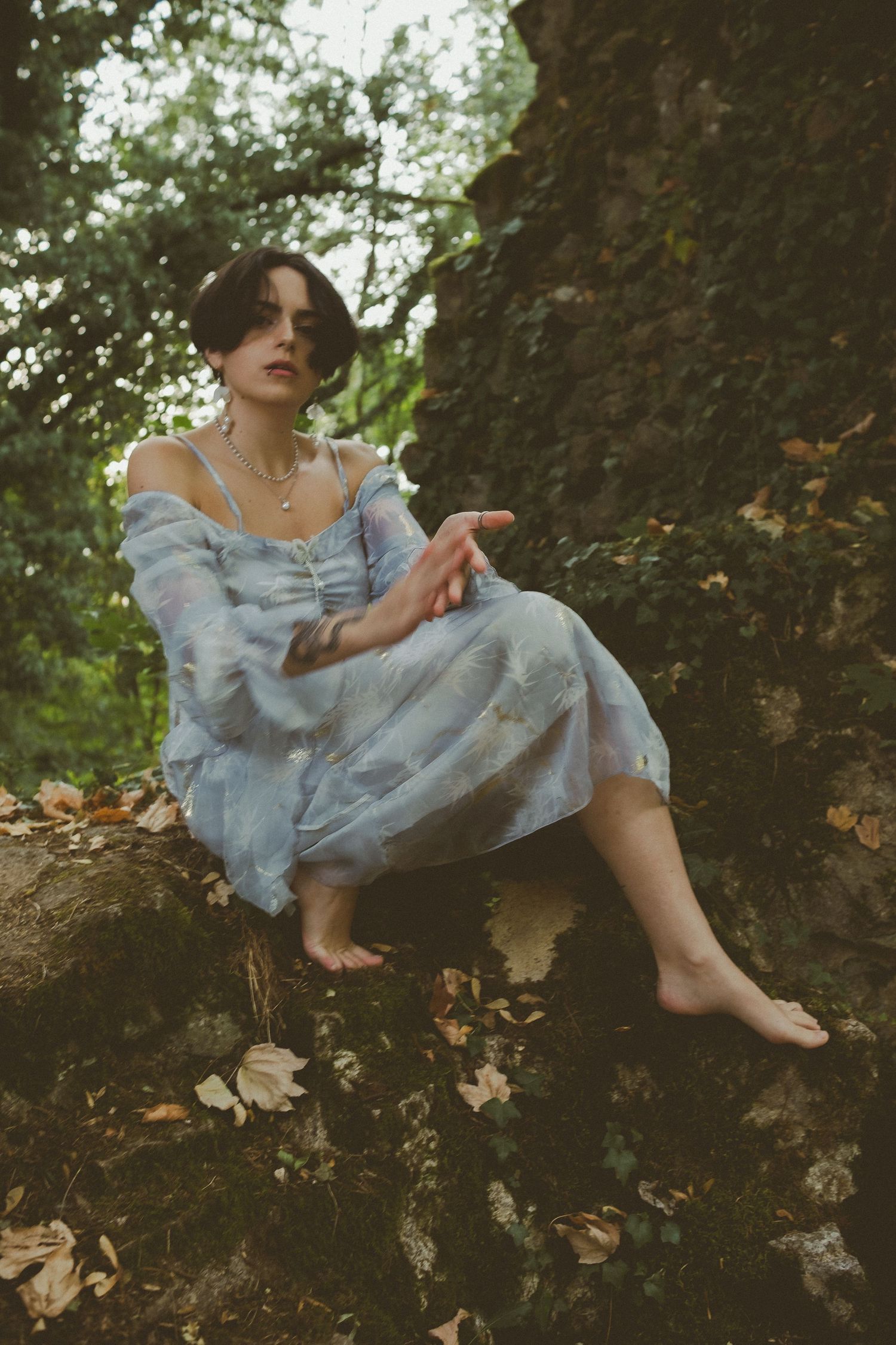 Young woman in flowy blue floral dress sits barefoot on mossy rocks surrounded by autumn leaves in a lush green forest.