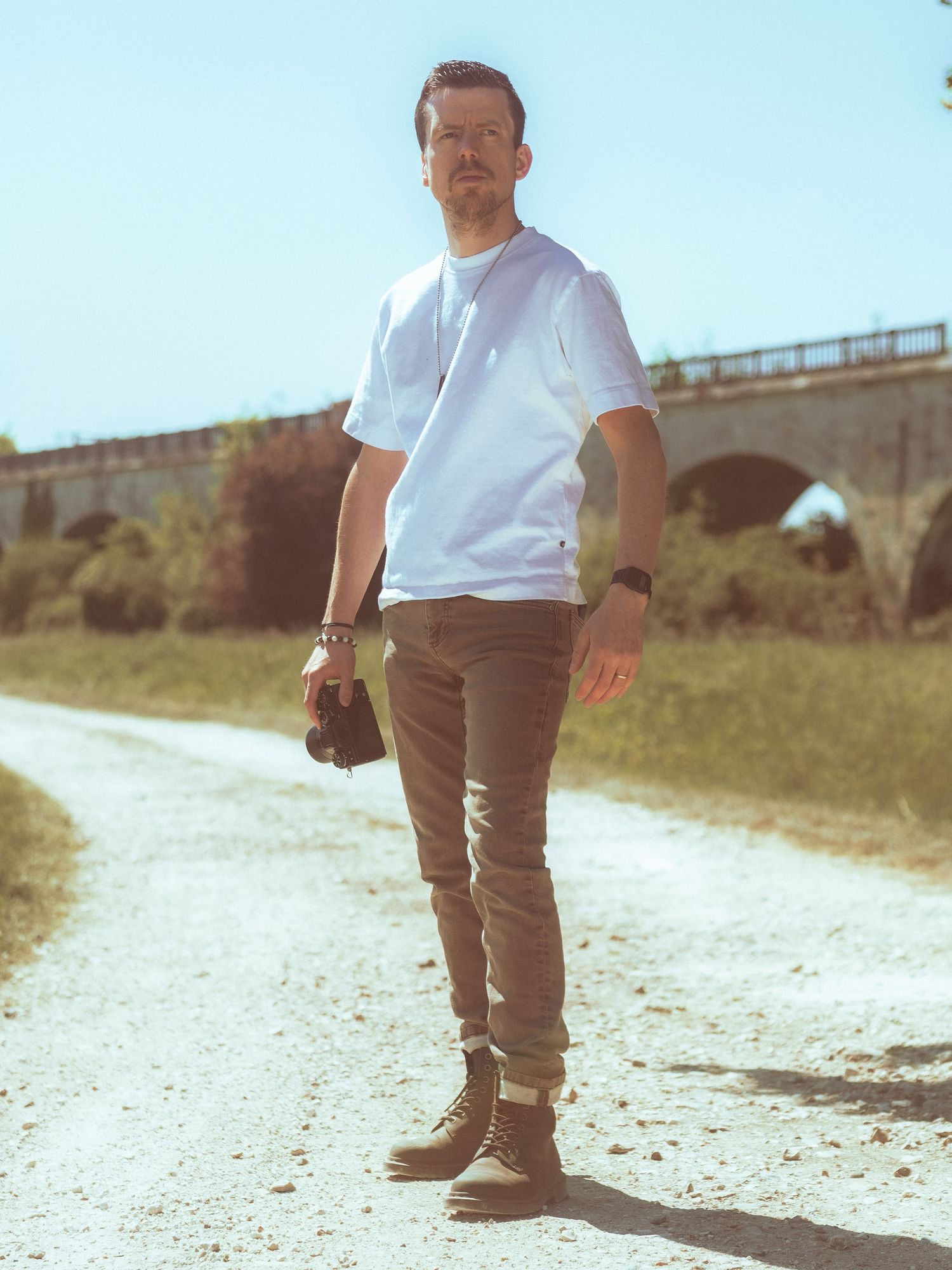 Photographe debout sur un chemin ensoleillé en terre près d'un pont en pierre, tenant un appareil photo.
