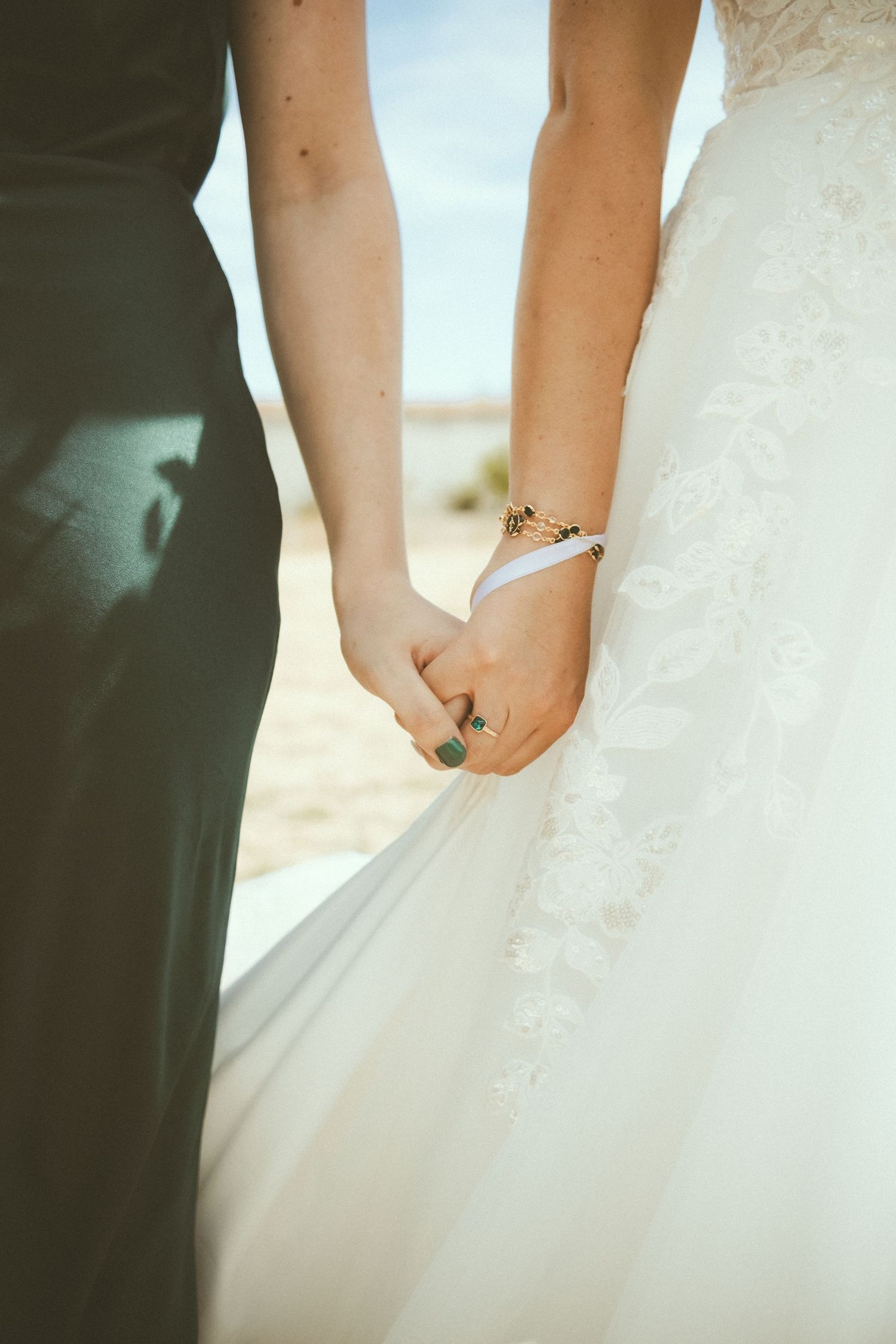 Two brides holding hands in wedding attire, one in white lace gown, outdoors in sunny desert setting.