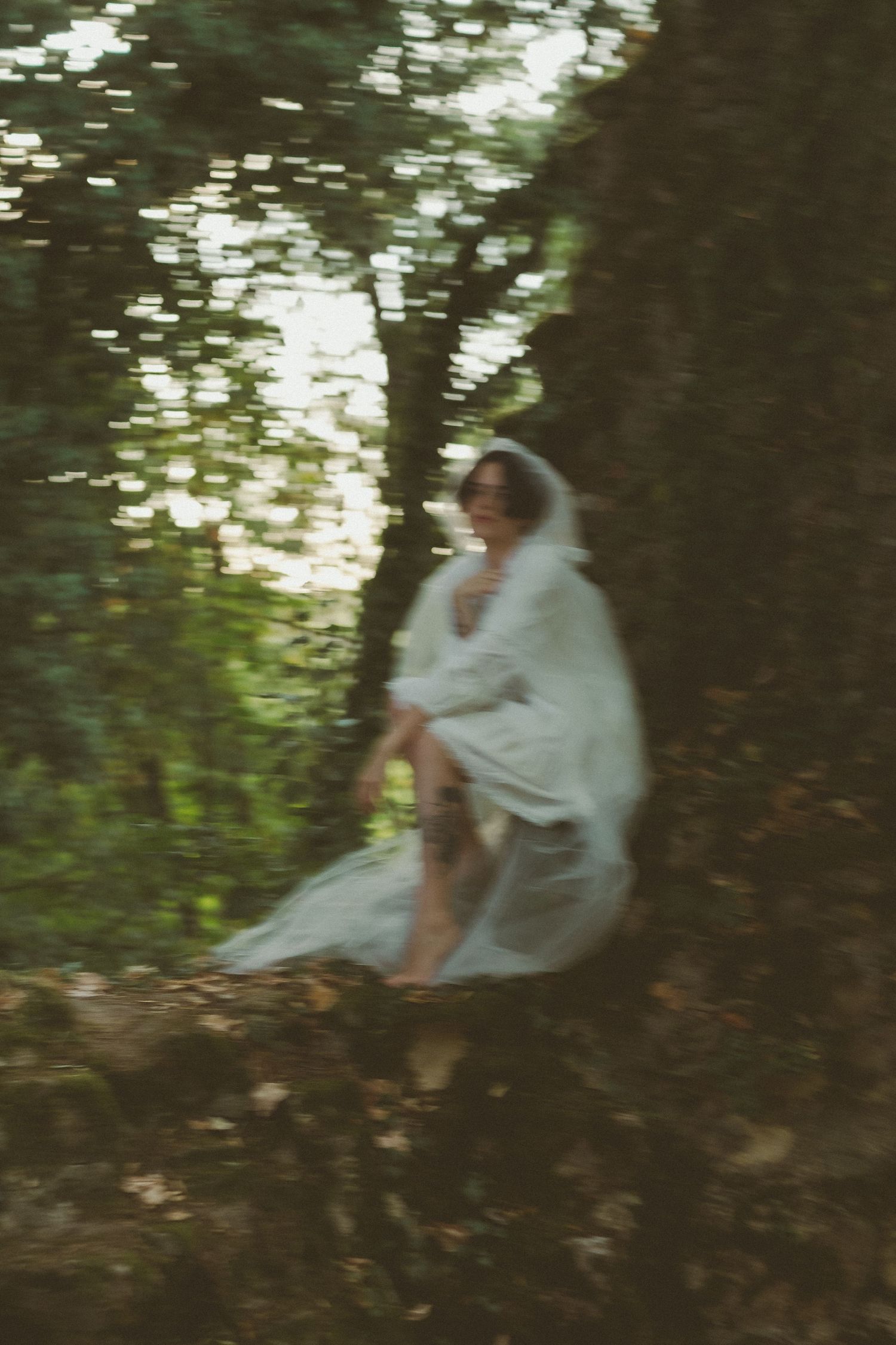 A blurred artistic shot of a woman in a white dress and veil seated among trees in nature.